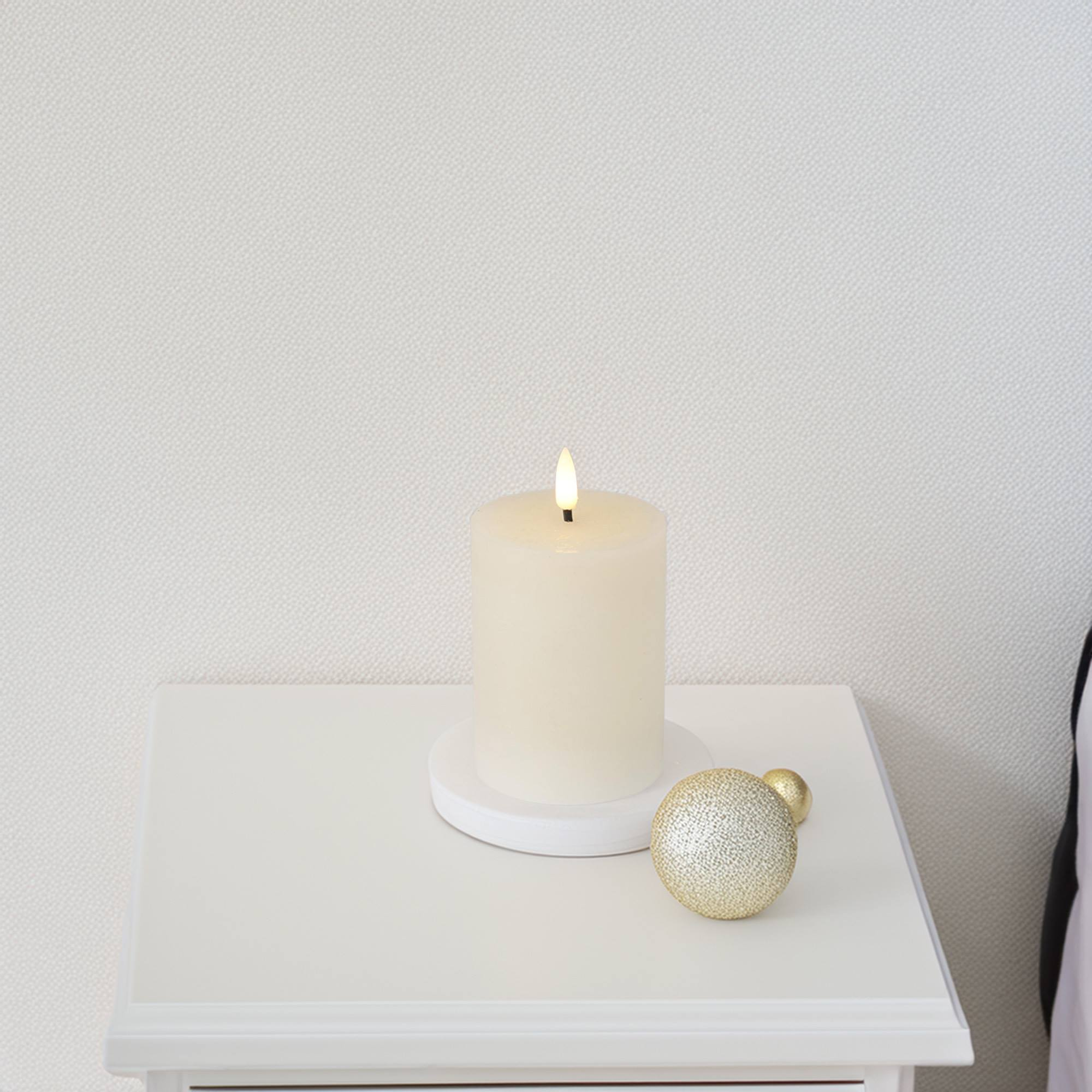 Una vela marfil encendida está sobre una mesa blanca con dos adornos dorados. El fondo es una pared blanca texturizada.