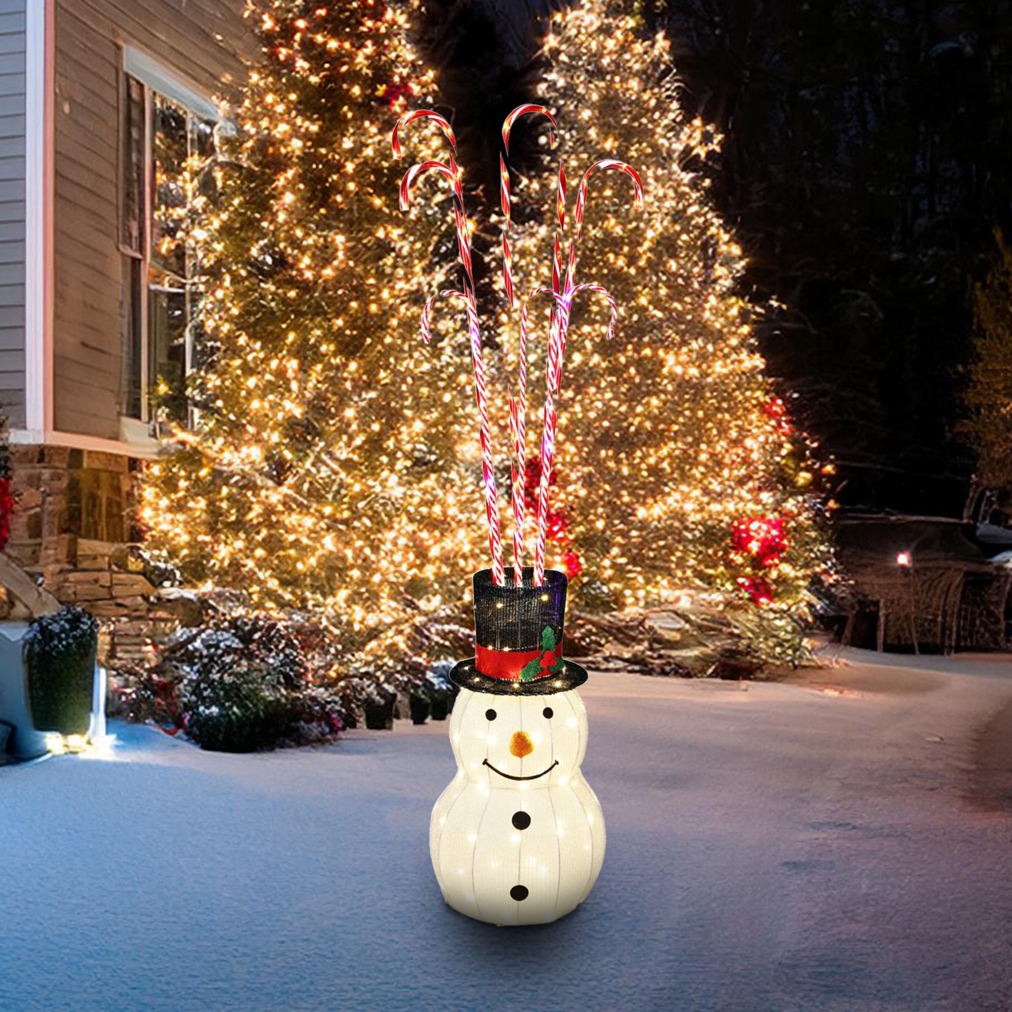 Un muñeco de nieve con sombrero y bastones de caramelo frente a árboles de Navidad iluminados. Nieve en el suelo.