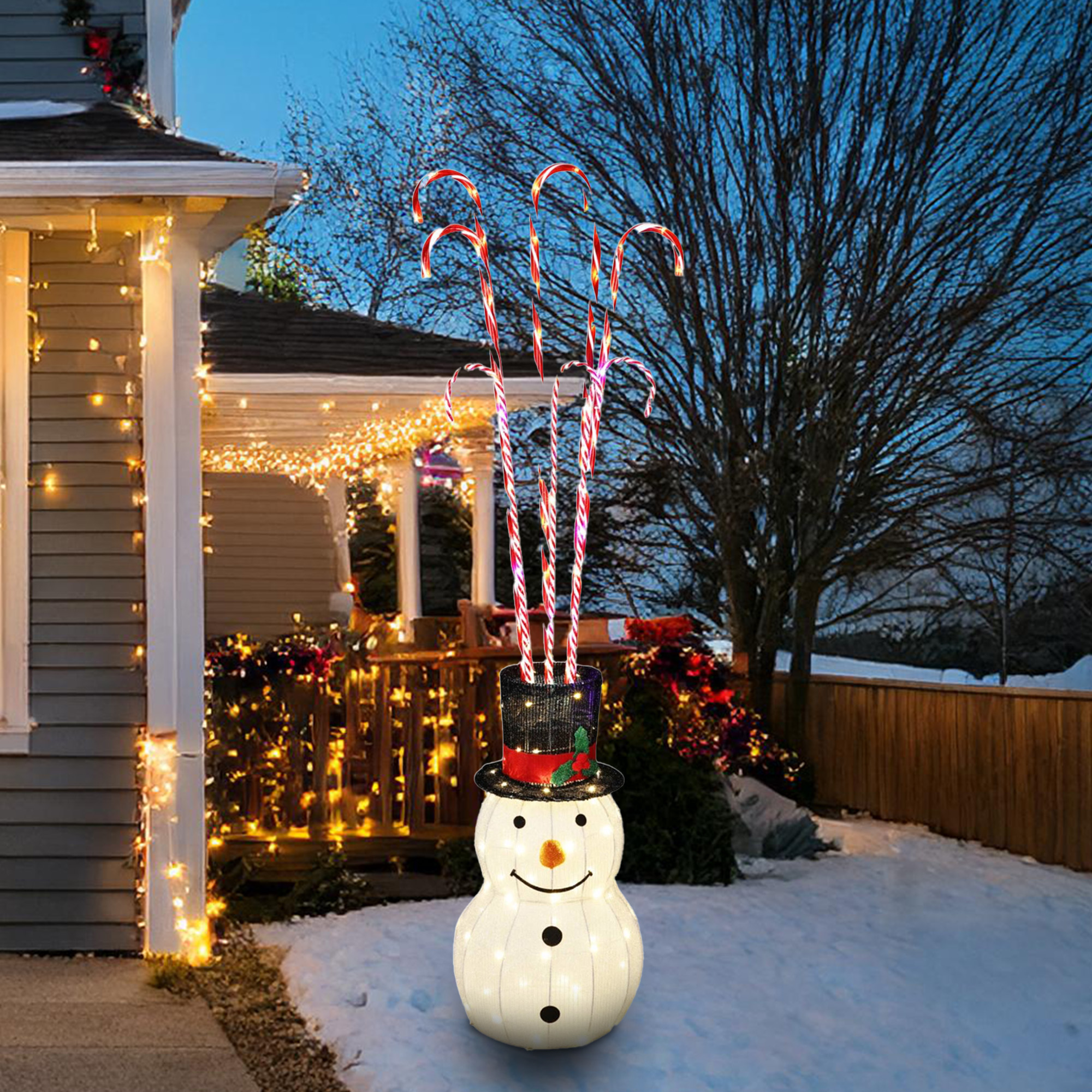 Un muñeco de nieve con sombrero sosteniendo bastones de caramelo. Una casa con luces en el fondo. Hay nieve en el suelo.