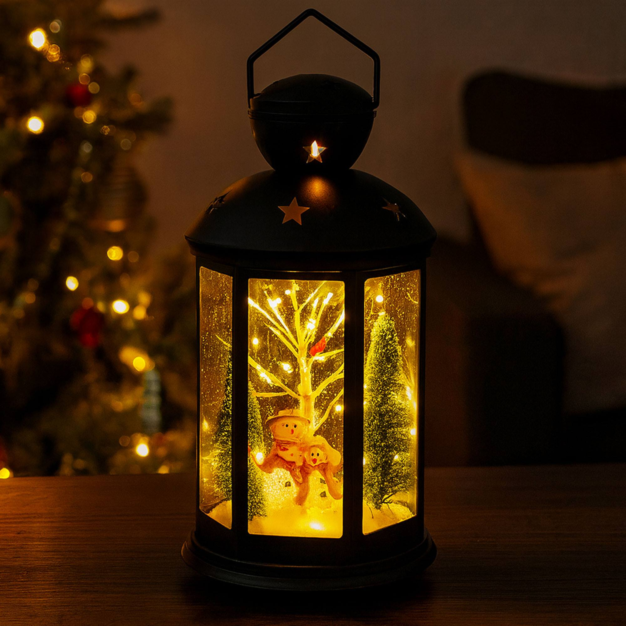 Una linterna navideña decorativa con muñecos de nieve y un árbol iluminado se coloca sobre una superficie de madera.