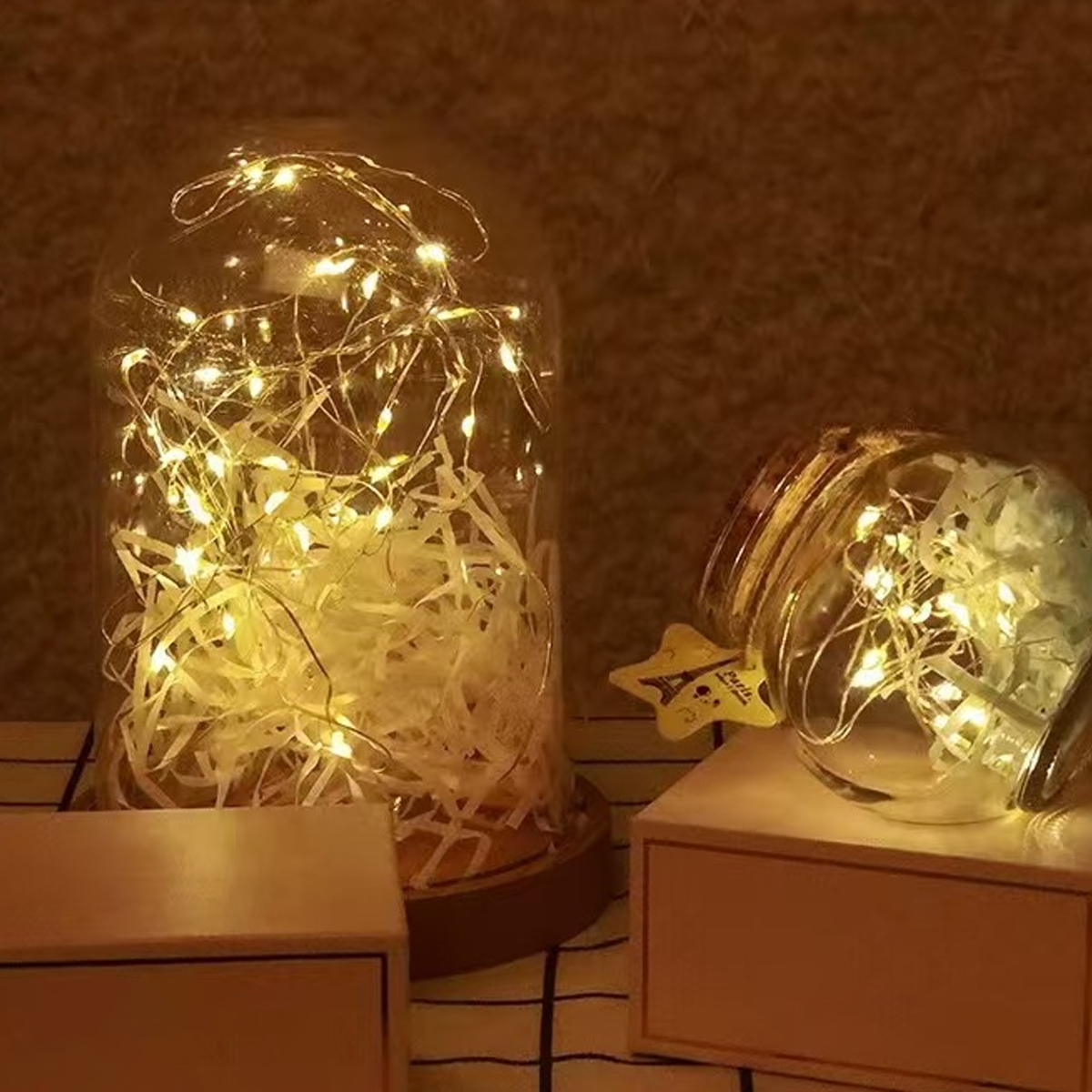 Zwei Glasbehälter mit Lichterketten. Einer aufrecht, mit Papier gefüllt. Der andere ist geneigt, auf einer Holzbox, mit einem Stern.