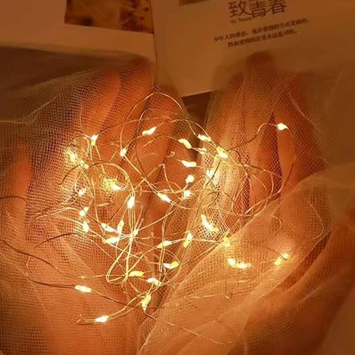 Hände halten ein Netz mit Lichterketten. Die warmen Lichter leuchten gegen das Netz. Im Hintergrund ist ein Buch zu sehen.