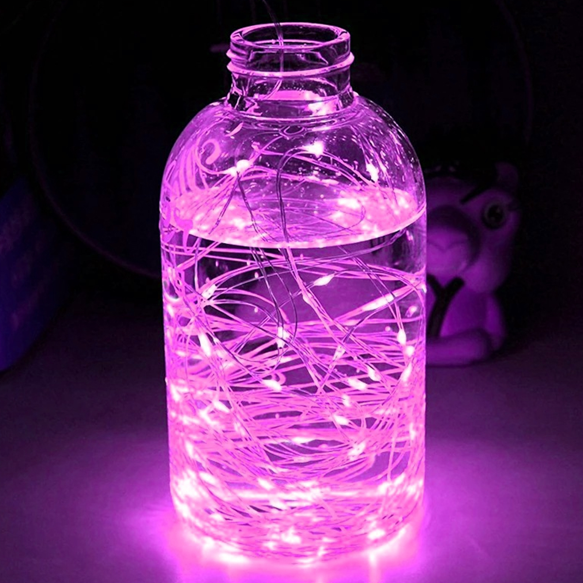 Ein klares Glasgefäß gefüllt mit Wasser und lila Feenlichtern. Im Hintergrund eine kleine lila Figur.