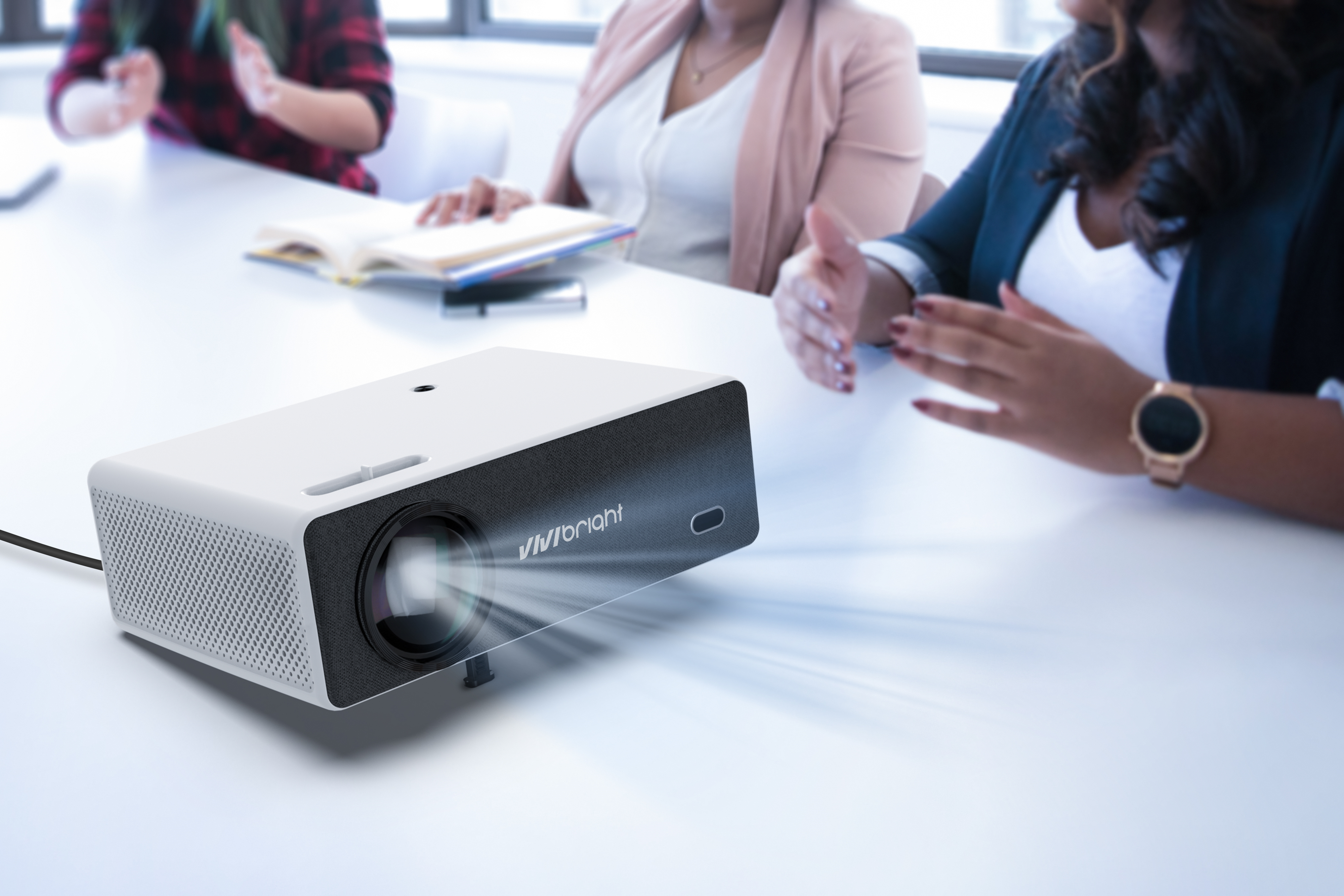 Un proyector blanco sobre una mesa blanca. Hay gente alrededor de la mesa. El proyector tiene la marca 'vivibright'.