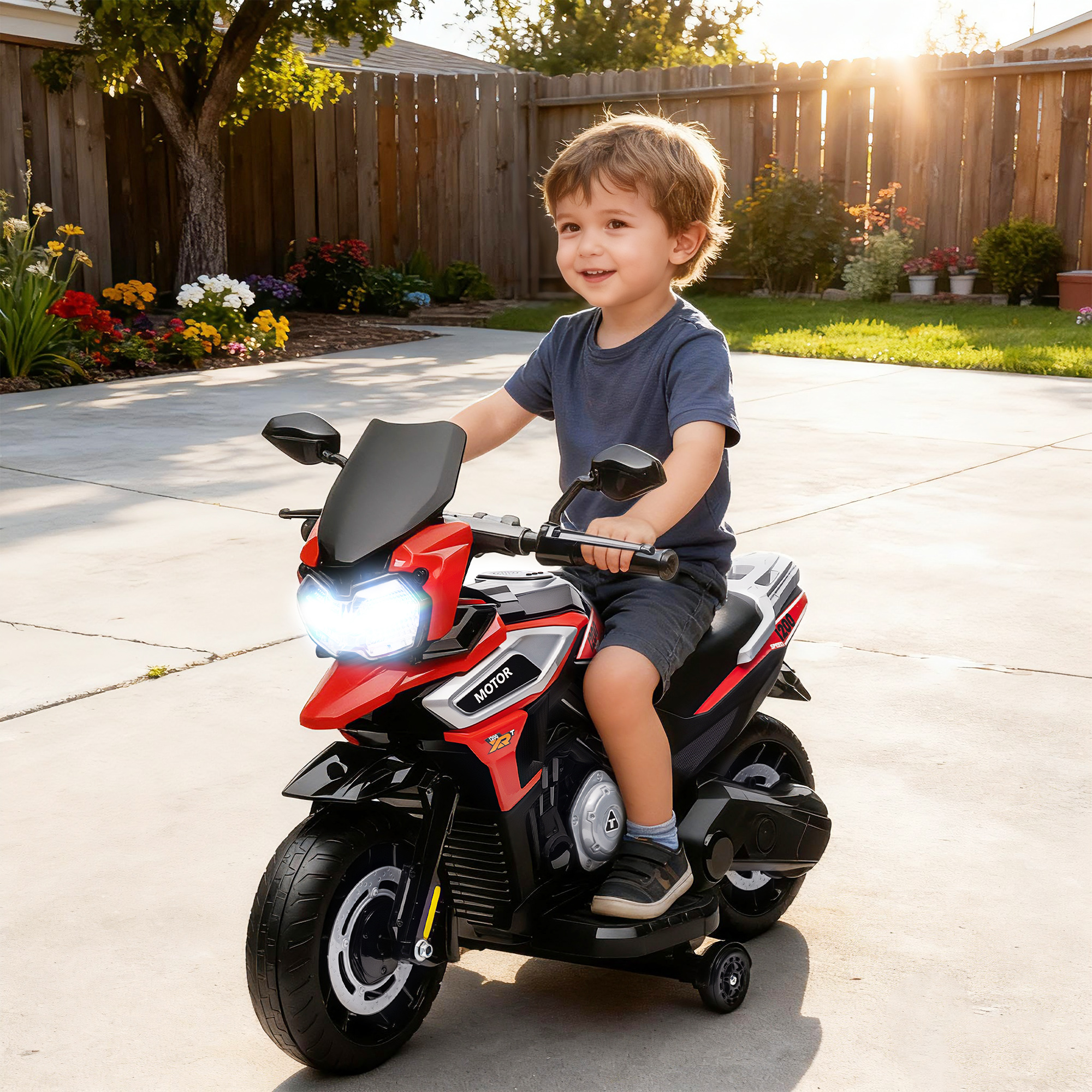 Un ragazzo sorridente guida una moto giocattolo rossa e nera all'aperto. Una staccionata di legno e fiori sullo sfondo.