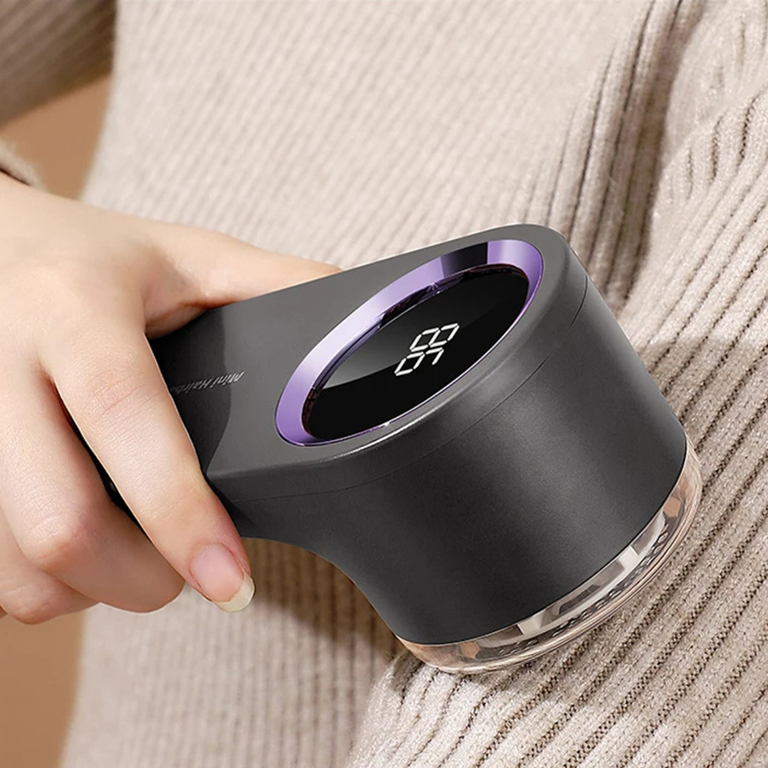 Eine Hand hält einen schwarzen Stoffrasierer mit einem lila Ring. Er liegt auf einem beigen Pullover. Der Rasierer zeigt eine Digitalanzeige.