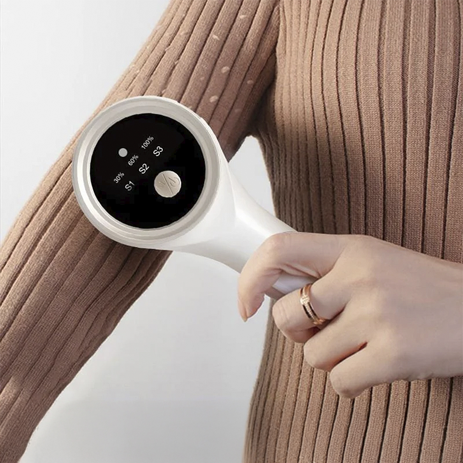 Eine Person hält ein weißes Gerät an ihrem Arm, trägt einen braunen Pullover und einen Ring.