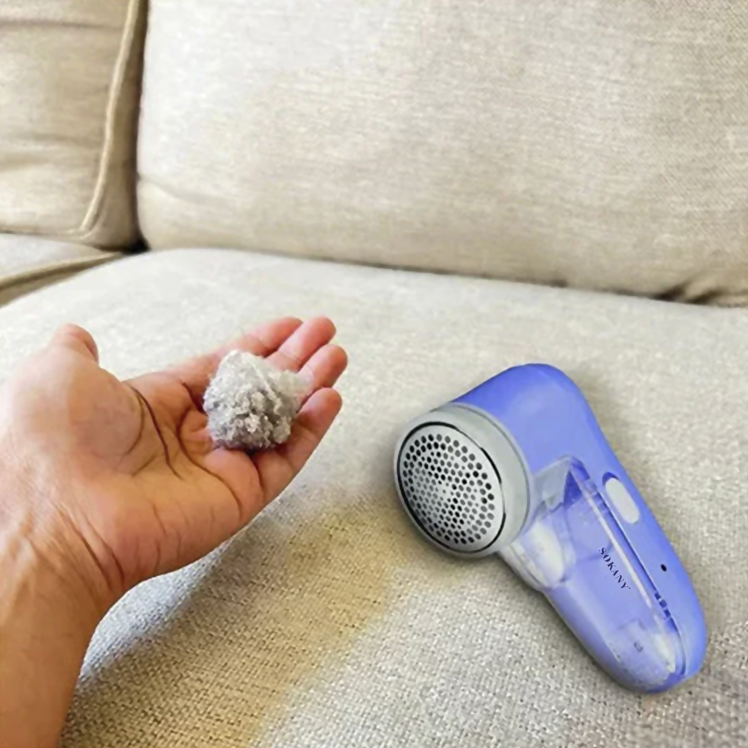 Eine Hand hält Fusseln von einem Pulloverrasierer. Ein blauer Pulloverrasierer liegt auf einer Couch. Die Couch ist beige.