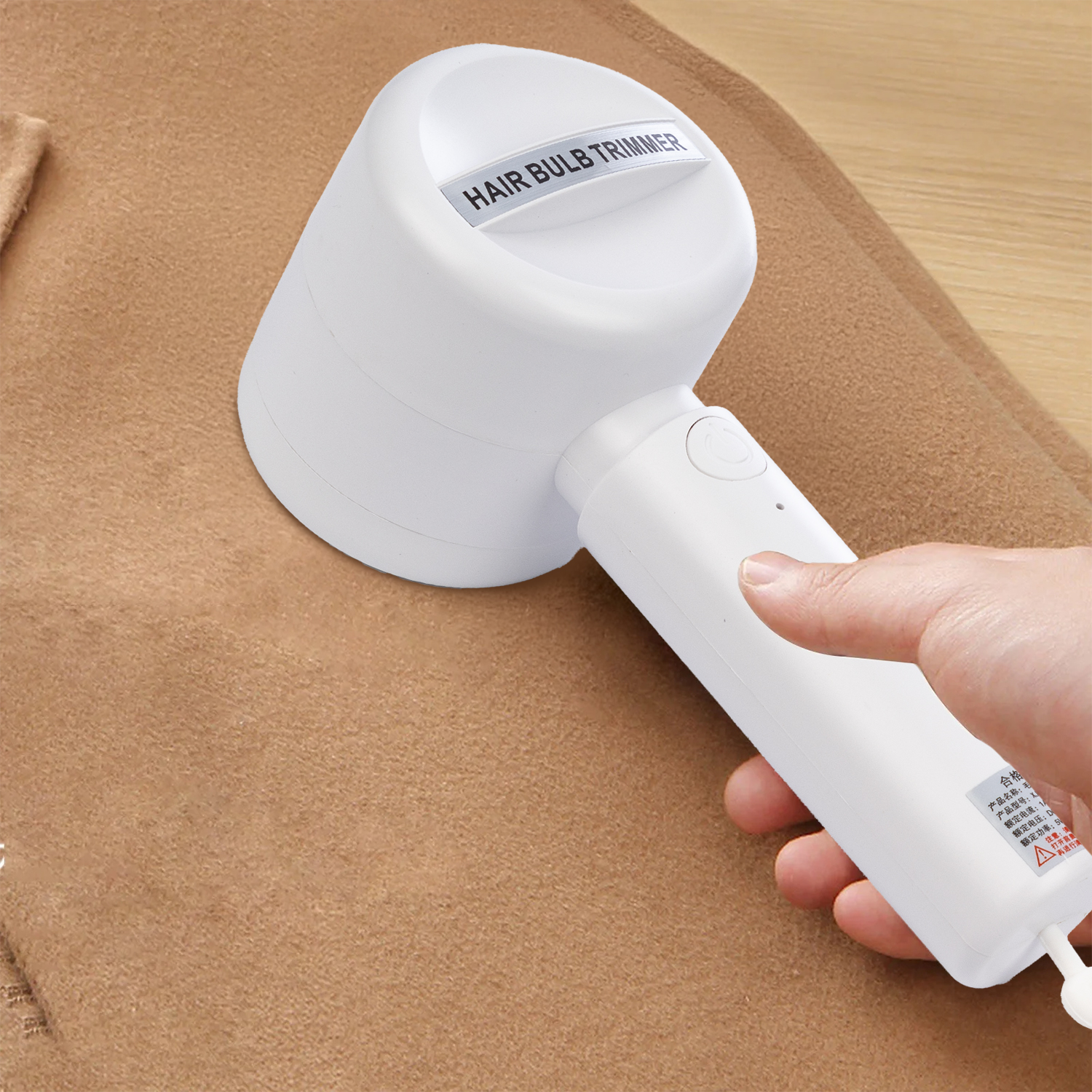 Ein weißer Haarzwiebeltrimmer wird von einer Hand gehalten. Er liegt auf einem braunen Stoff. Das Gerät ist mit 'HAIR BULB TRIMMER' beschriftet.