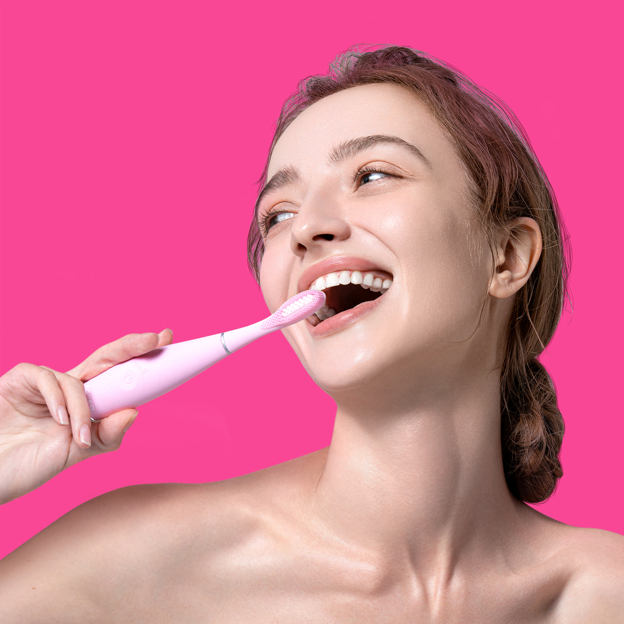 Una mujer se cepilla los dientes con un cepillo eléctrico rosa, sonriendo. Fondo rosa.