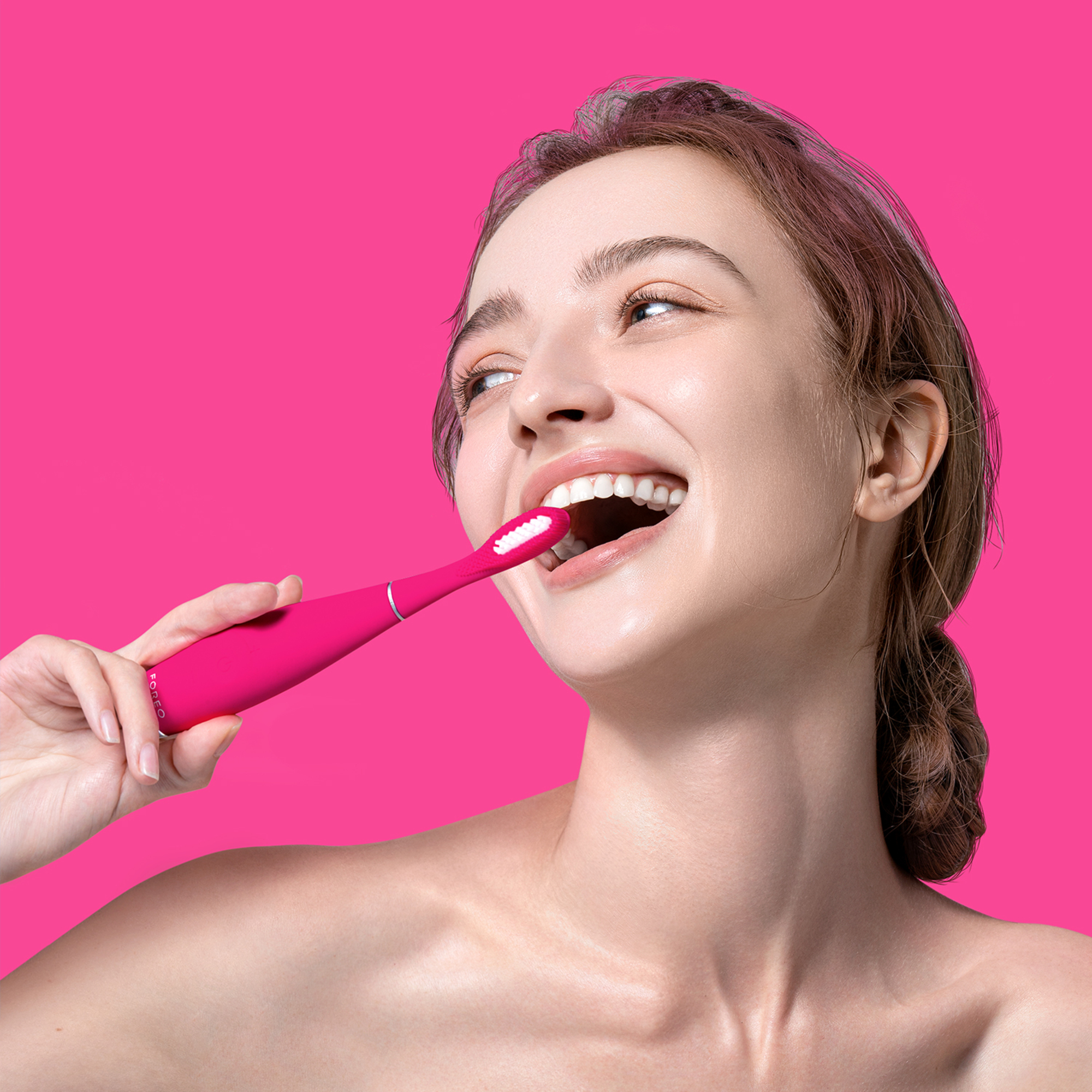 Una mujer se cepilla los dientes con un cepillo eléctrico rosa sobre un fondo rosa, sonriendo.