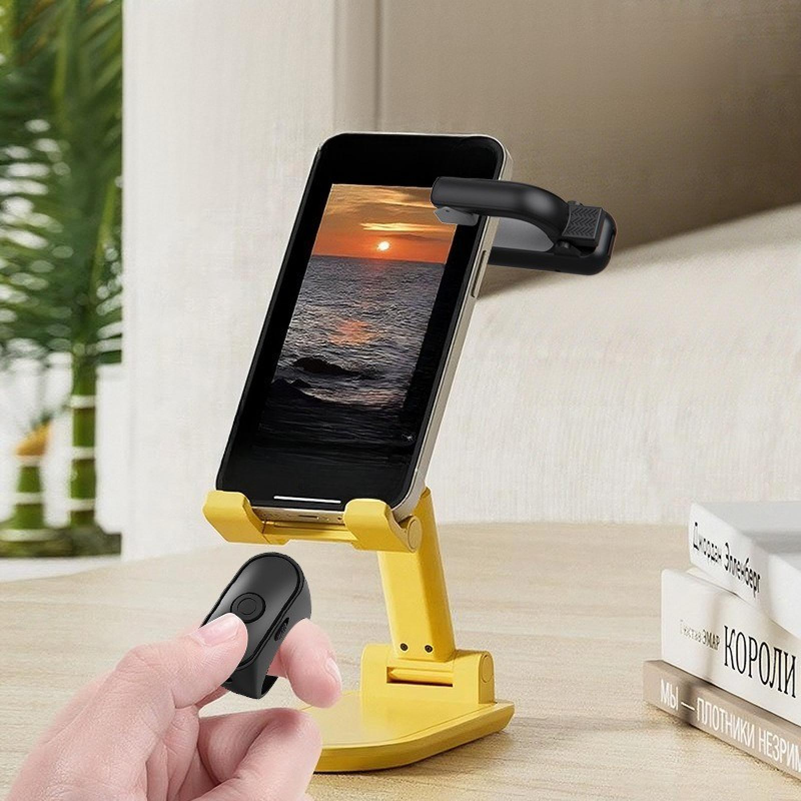 Ein Telefon auf einem gelben Ständer zeigt einen Sonnenuntergang über dem Meer. Eine Hand hält eine schwarze Fernbedienung. Bücher im Hintergrund.