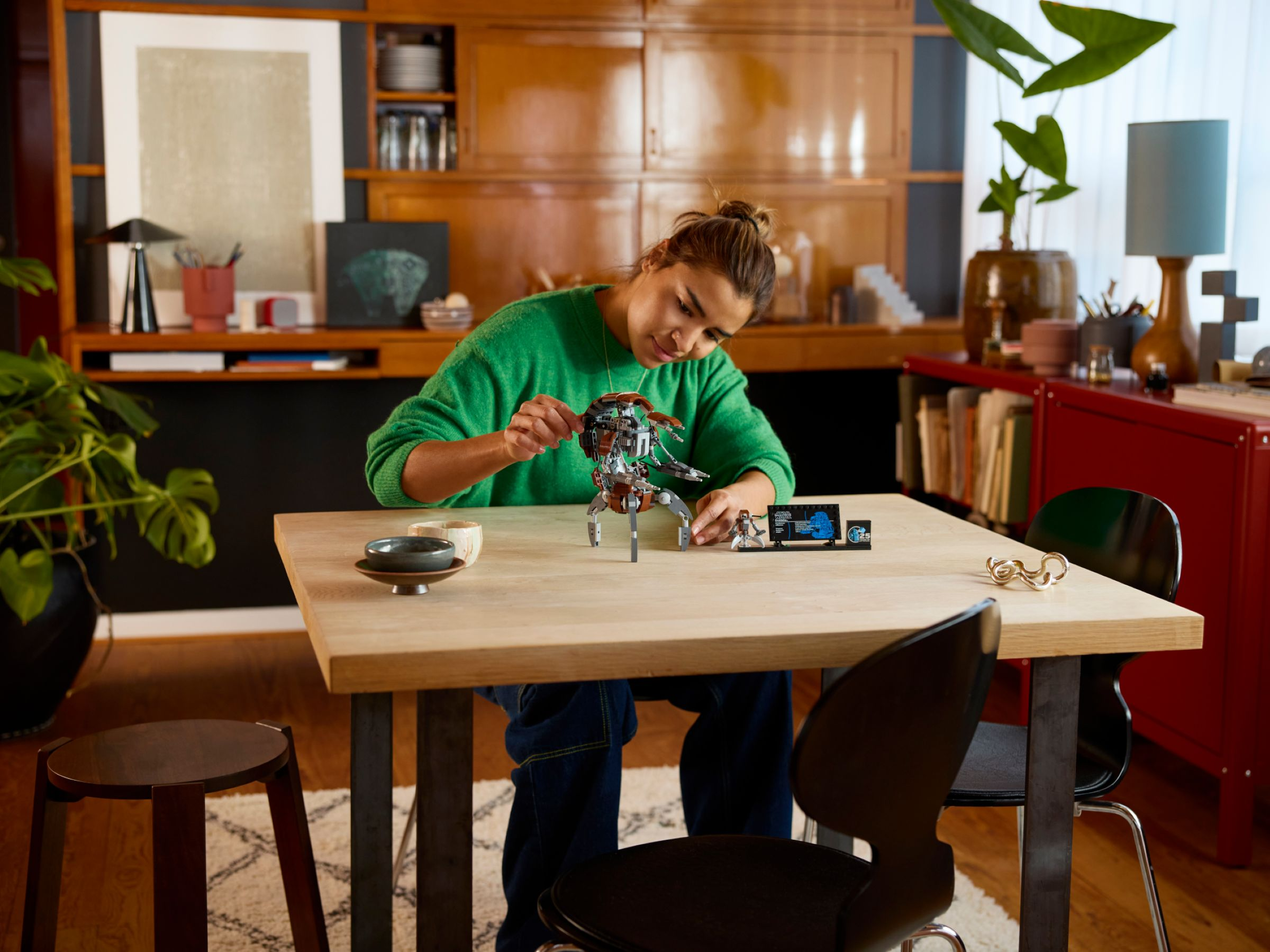 Eine Frau baut ein Roboterspielzeug auf einem Holztisch. Der Hintergrund umfasst einen Schrank, Pflanzen und einen roten Schrank.