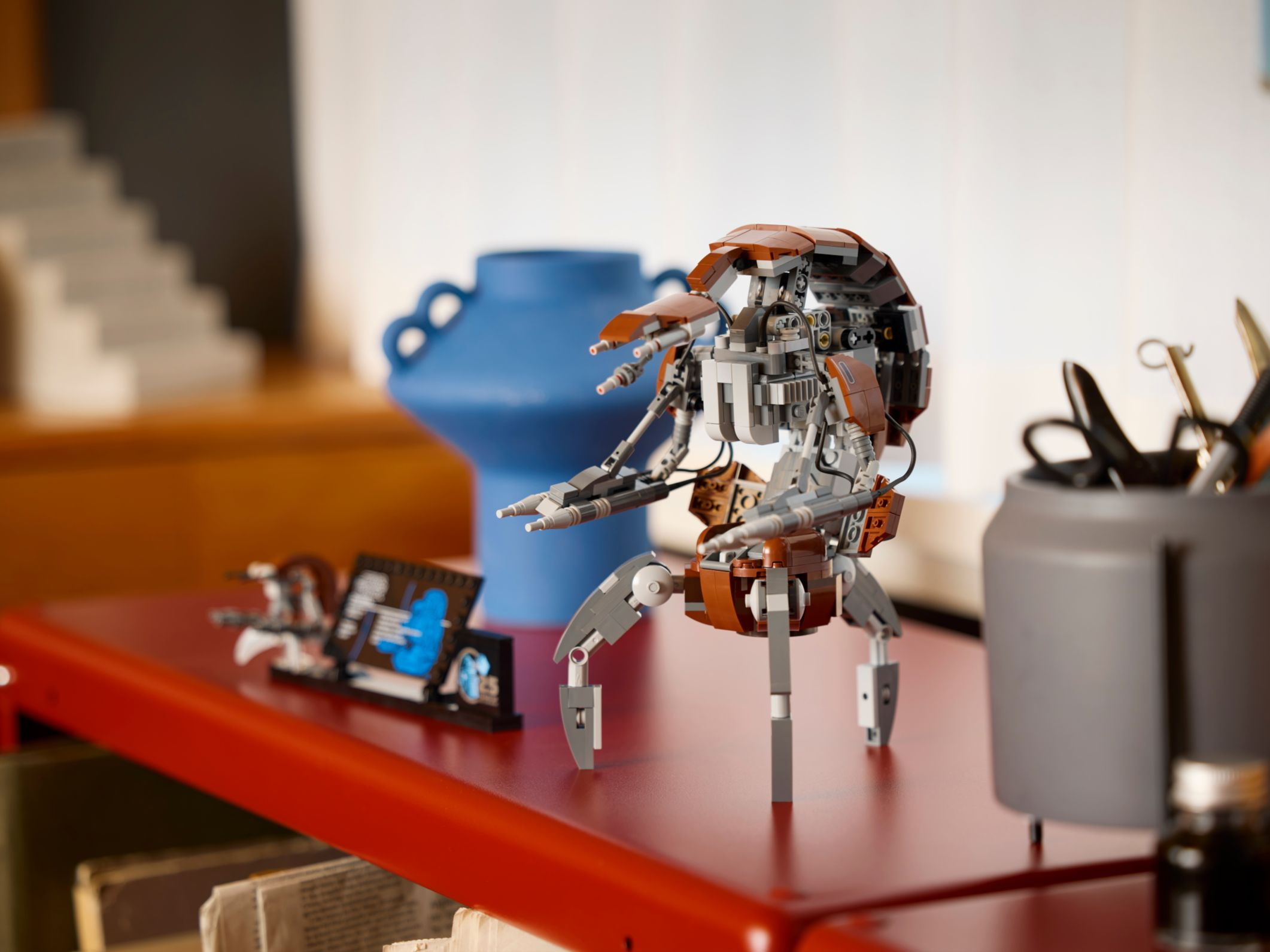 Ein Lego-Roboter steht auf einer roten Oberfläche. Eine blaue Vase, eine Schere und andere Gegenstände sind im Hintergrund.