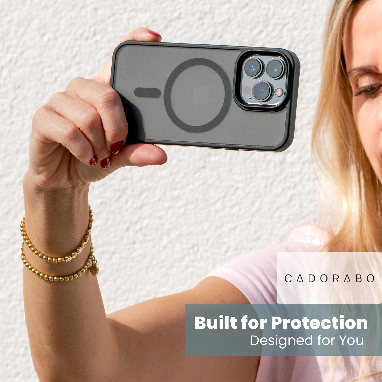 Eine Frau hält ein Telefon mit Hülle. Zwei goldene Armbänder sind an ihrem Handgelenk. Der Hintergrund ist eine weiße Wand.