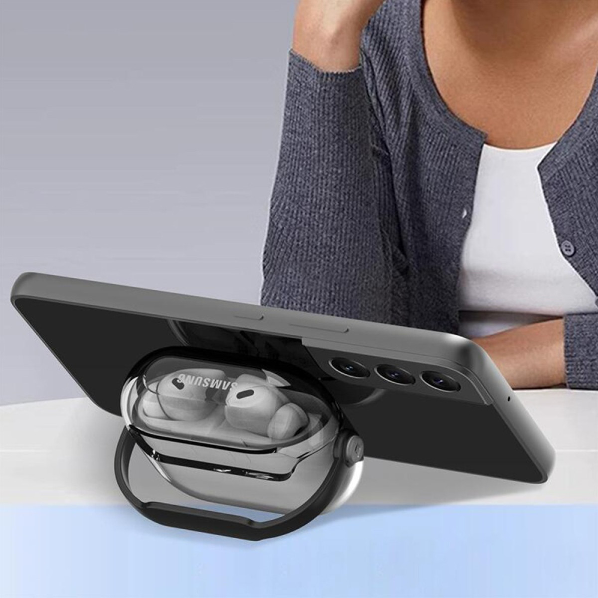 A phone with a case is propped up by a clear stand that holds wireless earbuds. A person is in the blurred background.