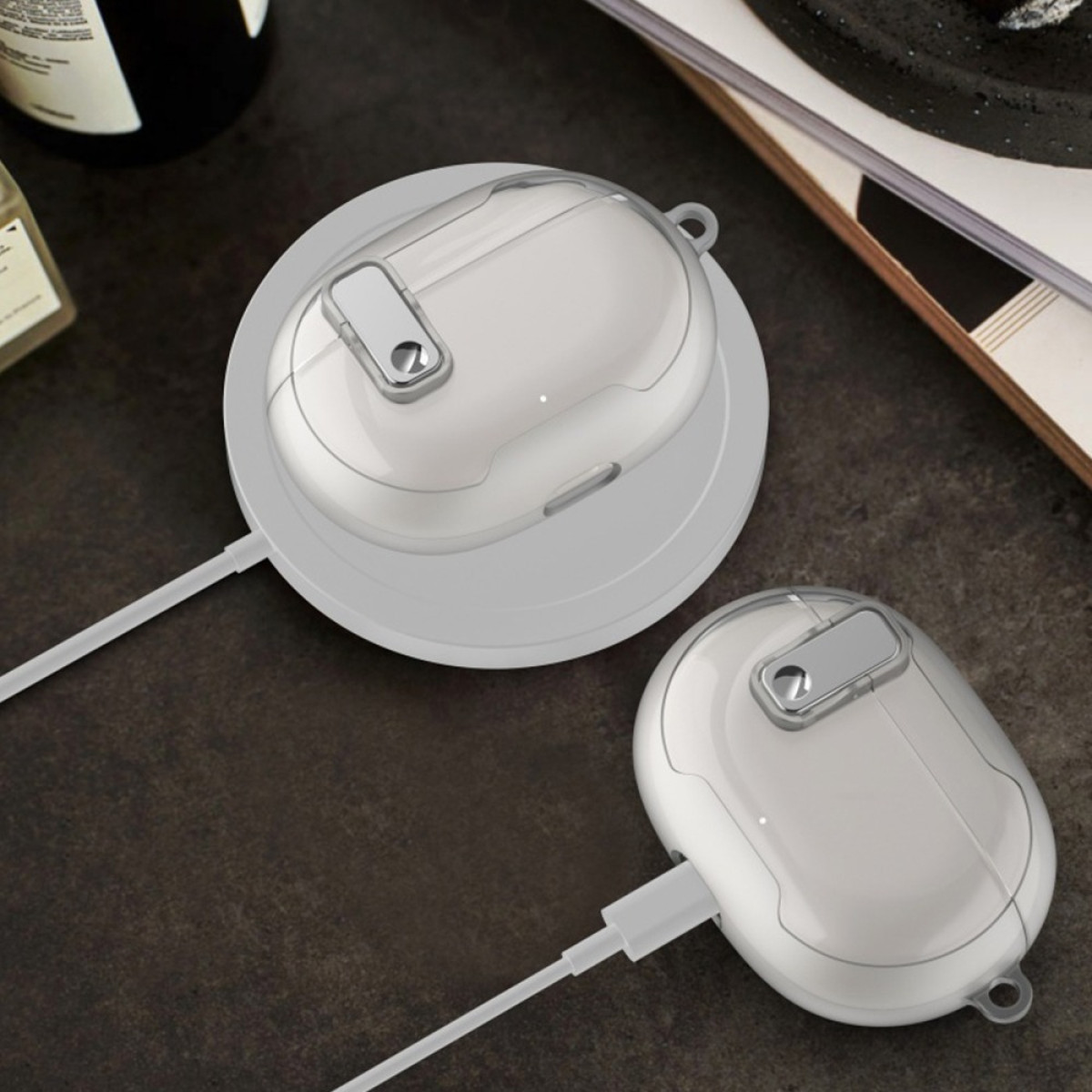 Two white earbud cases, one on a charging base, both with clear tops. Cables and parts of other objects are visible.