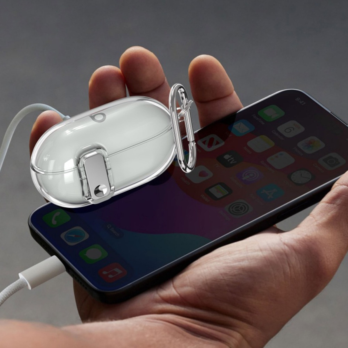 A hand holds a phone with a white earbud case. The phone is charging. The background is a blurry gray.