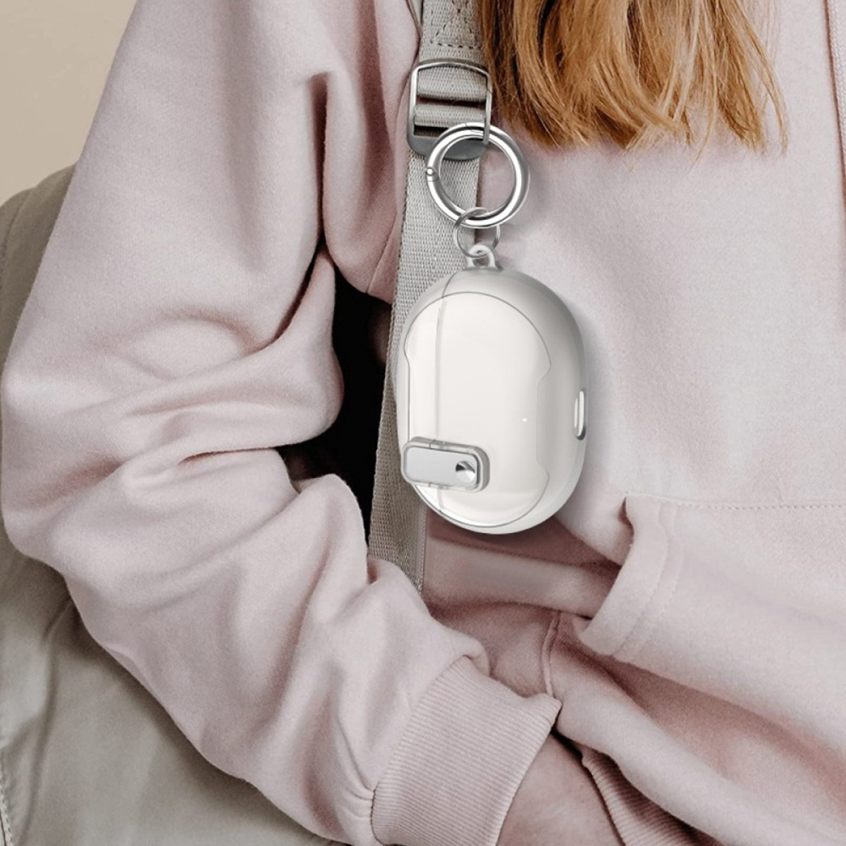 A white device is attached to a backpack strap via a ring. It hangs against a pink hoodie, and blond hair is visible.