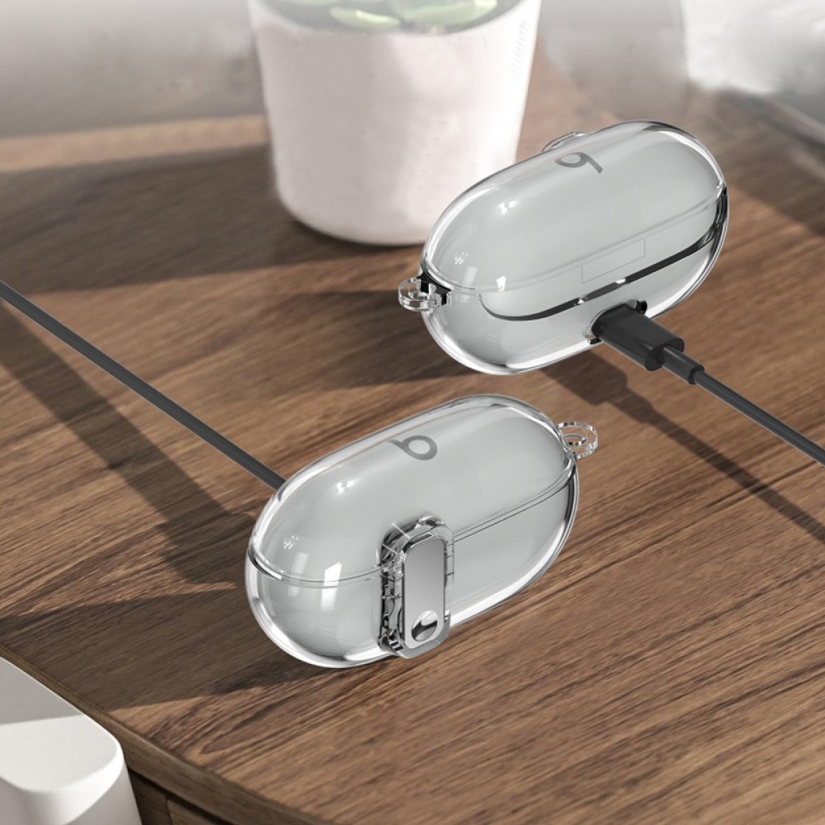 Two clear cases with earbuds on a wooden surface. One is charging with a black cable. A white pot is in the background.
