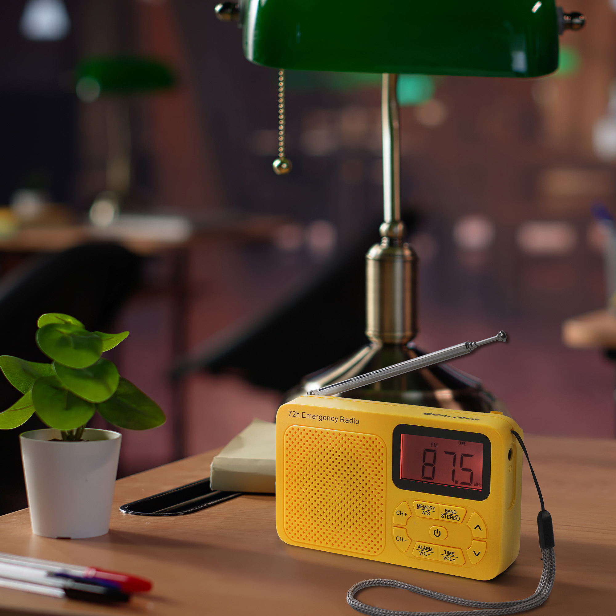 Yellow emergency radio with a screen displaying 87.5 MHz. A green lamp and a small plant are on the wooden table.