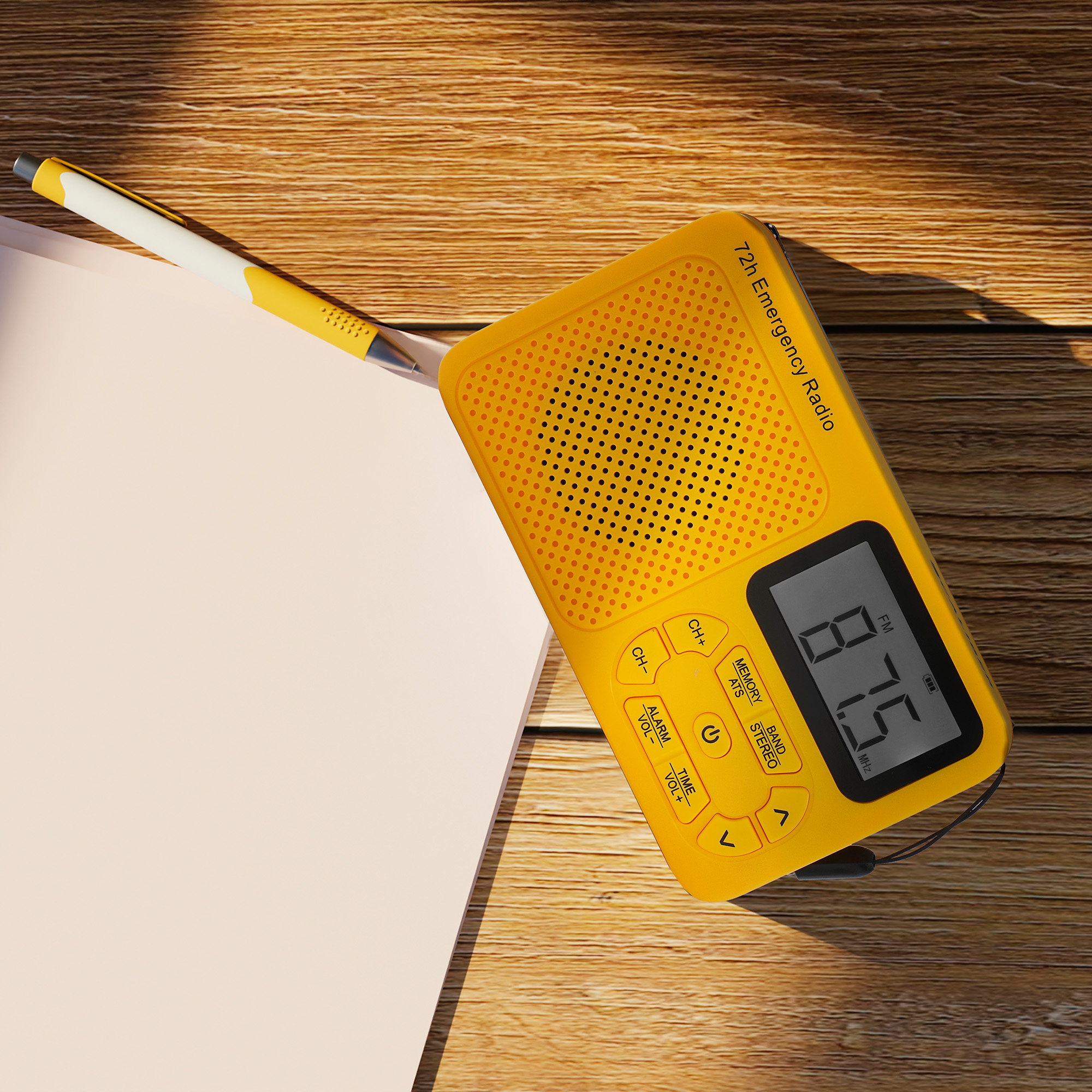 Yellow emergency radio with a digital display, pen, and notebook on a wooden surface. The radio is angled.