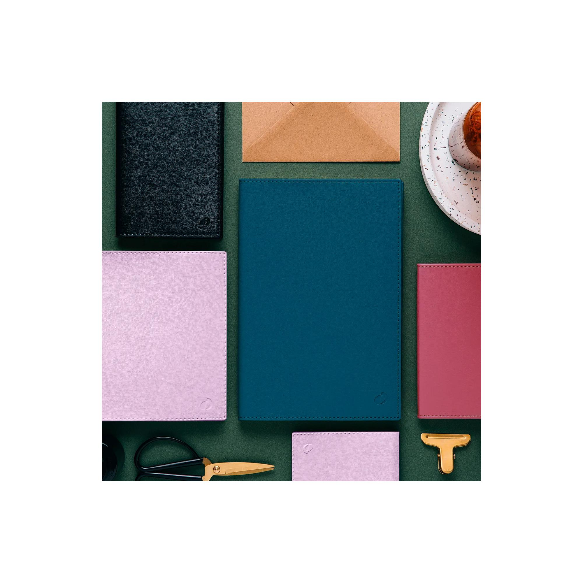 Vista cenital de cuadernos de varios colores, un sobre, tijeras, una taza de té y un clip en forma de T sobre una superficie verde.