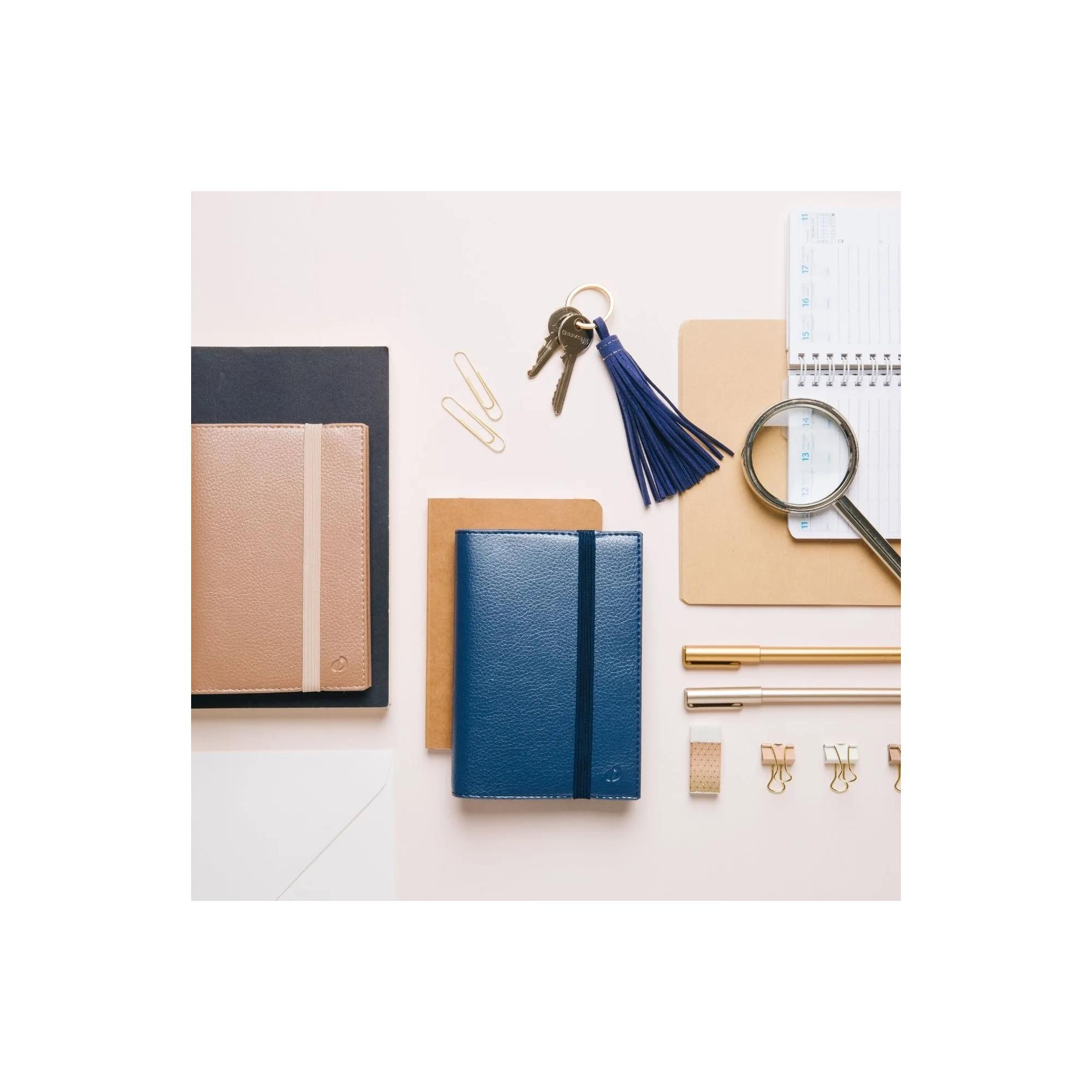 Schreibtischszene mit Notizbüchern, Schlüsseln, Kalender, Stiften, Büroklammern und einer Lupe auf rosa Oberfläche.