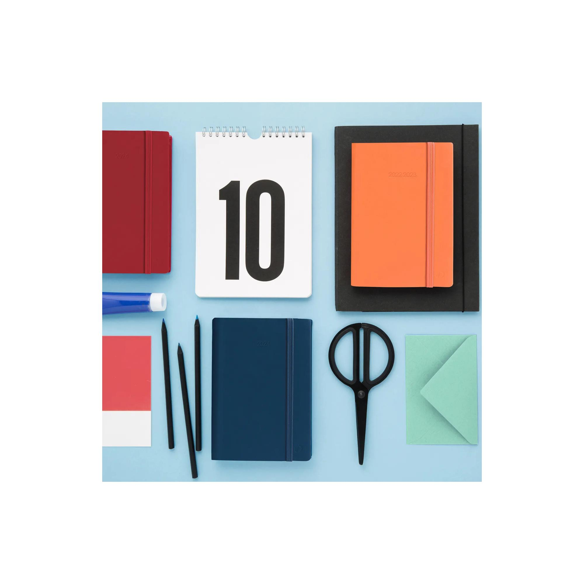 Vista cenital de útiles de escritorio. Incluye cuadernos, lápices, tijeras, un calendario y un sobre, todo sobre una superficie azul.