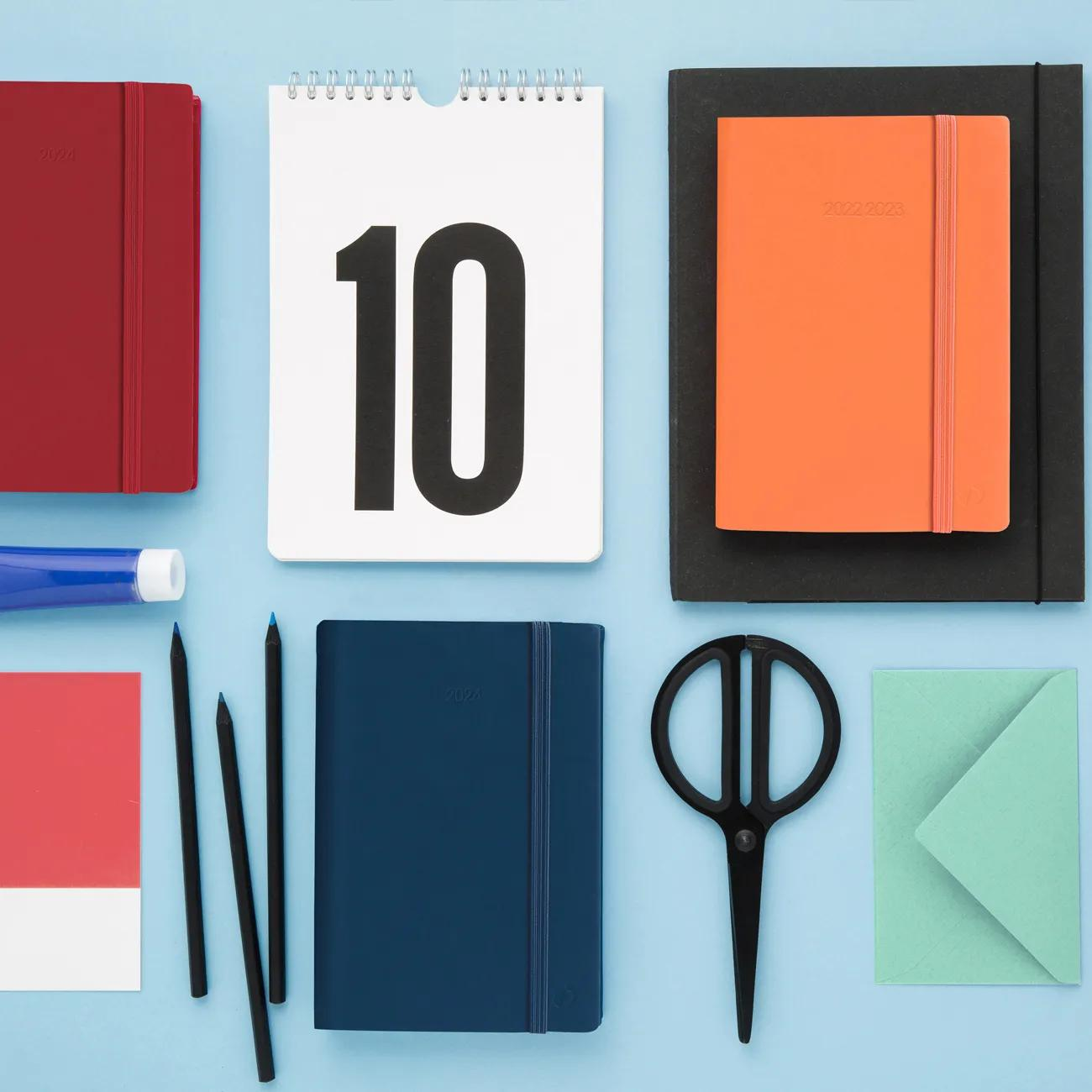 Una vista superior de útiles de oficina, incluyendo un calendario, cuadernos, lápices, tijeras y un sobre sobre fondo azul.