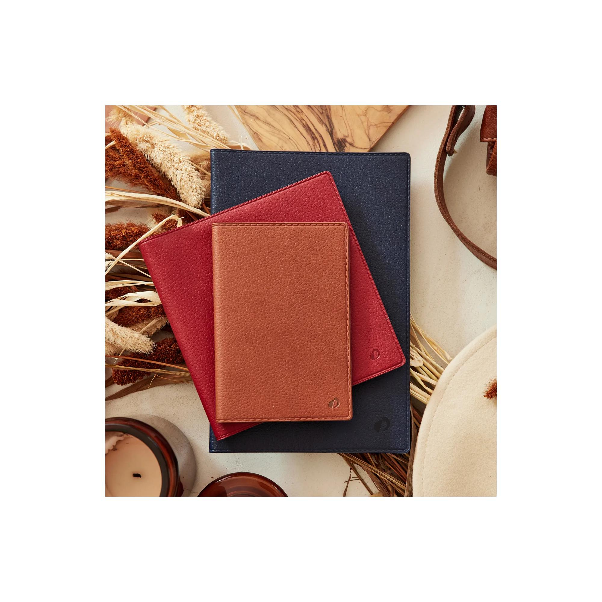 Tres carteras de cuero, rojas, marrones y azul marino, apiladas sobre una superficie beige con plantas secas y una vela.