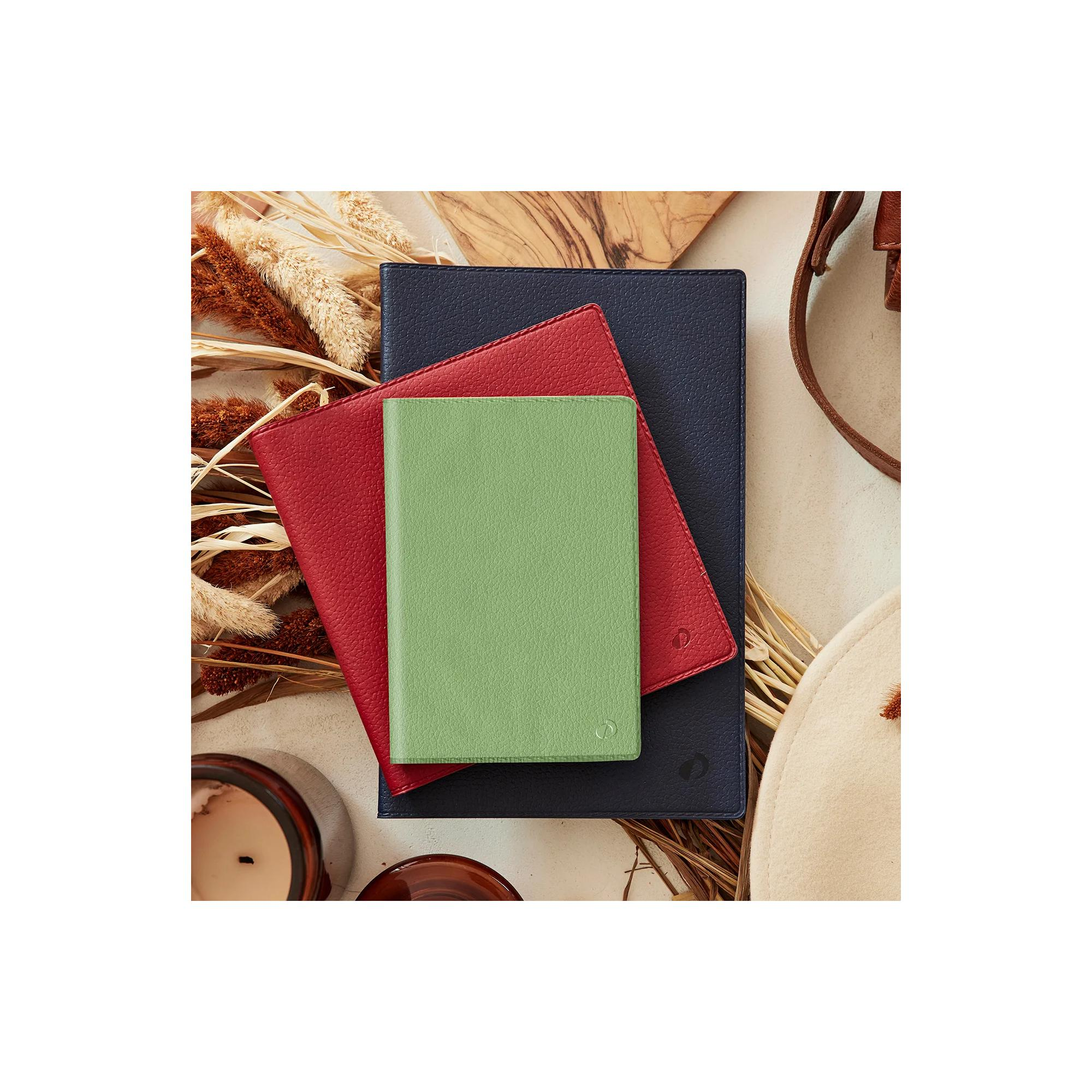 Tres cuadernos de cuero apilados: azul marino, rojo y verde. Plantas secas, una vela y un sombrero en el fondo.