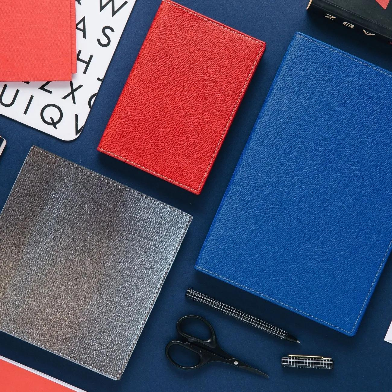 Vista cenital de cuadernos rojos, plateados y azules, con tijeras, un bolígrafo y un libro sobre una superficie azul oscuro.