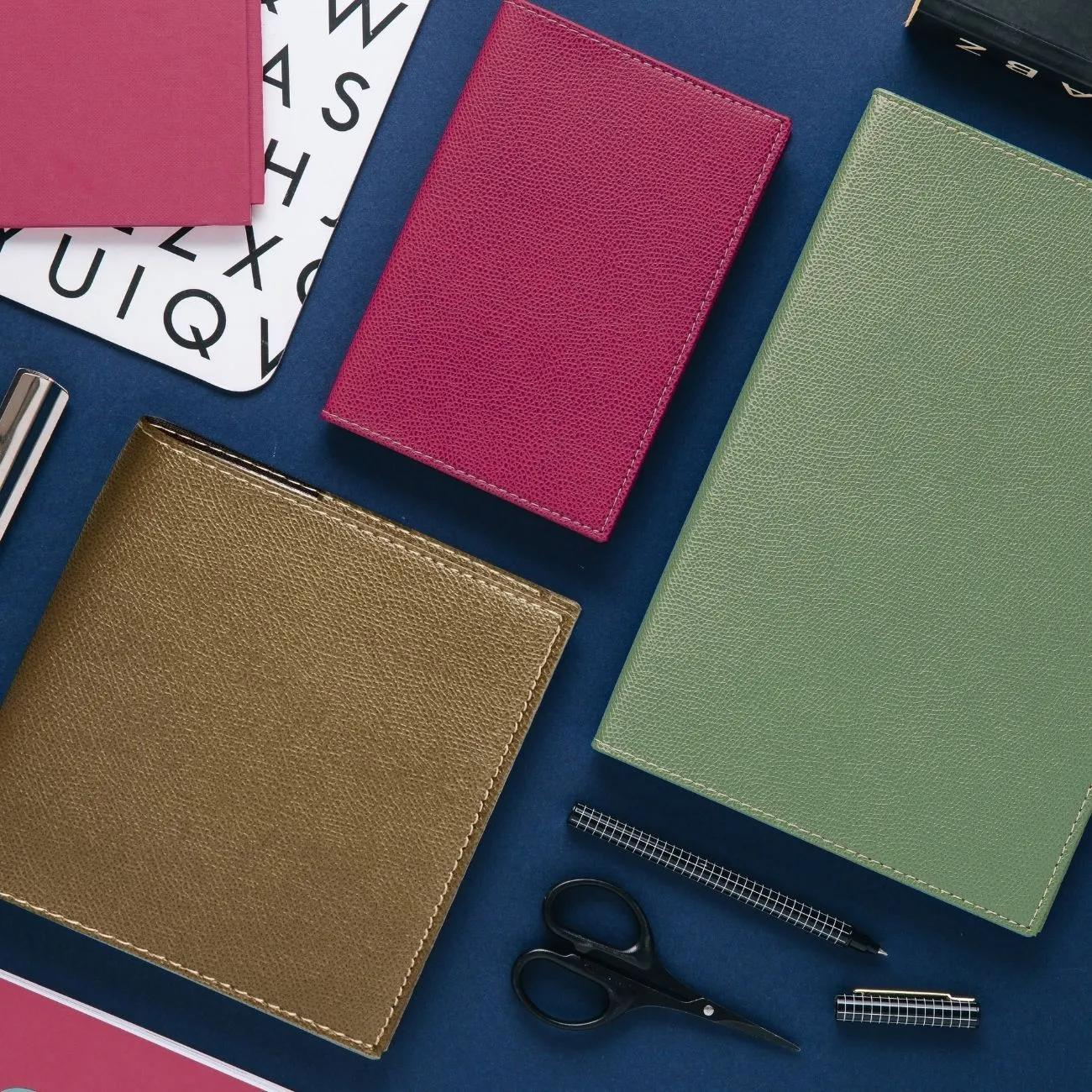 Toma cenital de cuadernos, bolígrafo, tijeras y tarjetas alfabéticas sobre superficie azul. Colores: borgoña, verde y dorado.