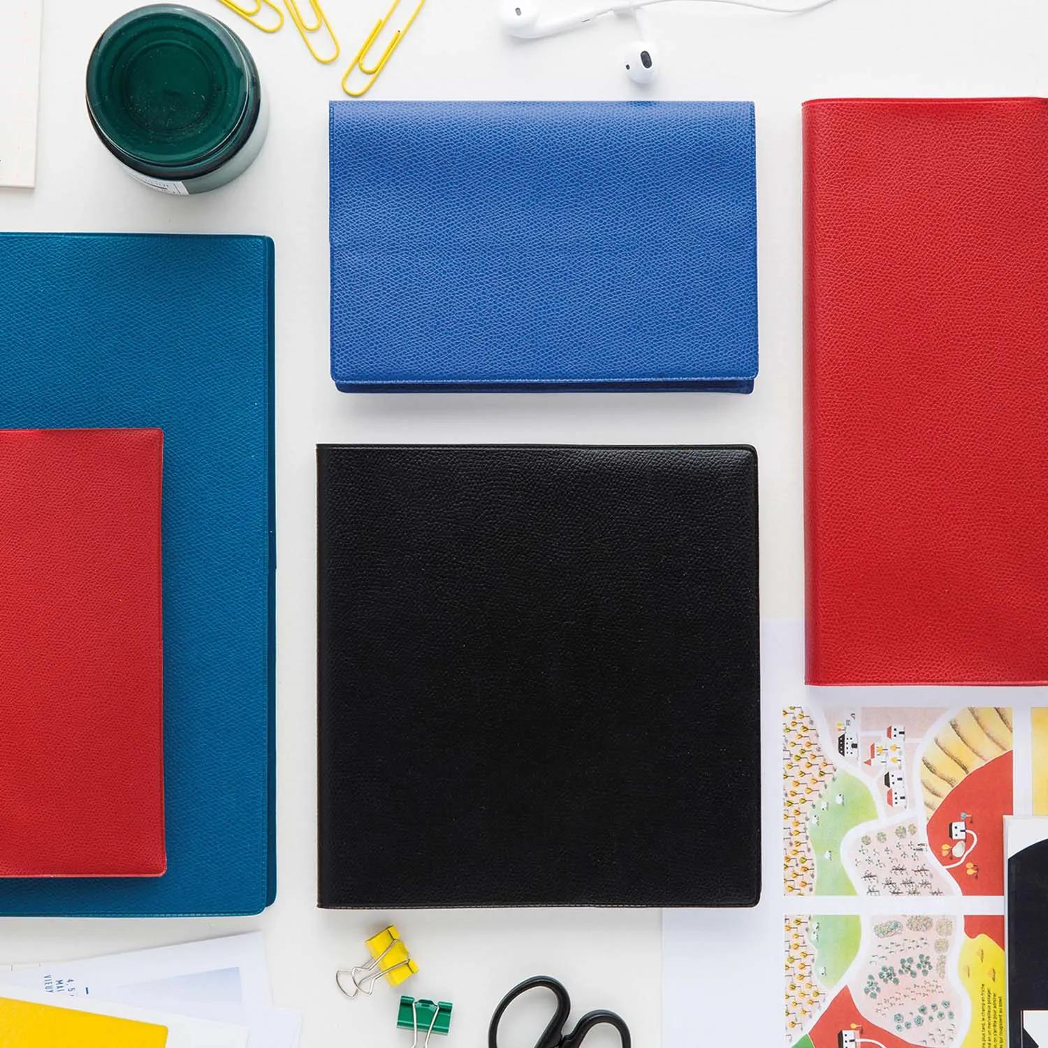 Toma cenital. Varios cuadernos de colores (azul, rojo, negro) con artículos de papelería como clips, tijeras y un frasco verde sobre una superficie blanca.