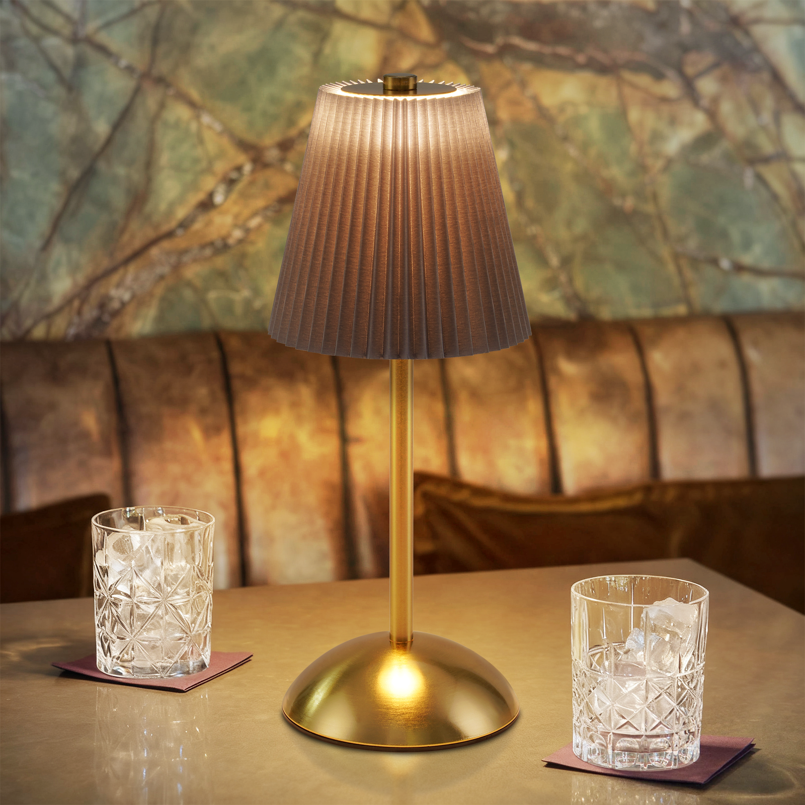 Una lampada in ottone illuminata con paralume plissettato è su un tavolo con due bicchieri di ghiaccio. Lo sfondo ha un muro marmorizzato.