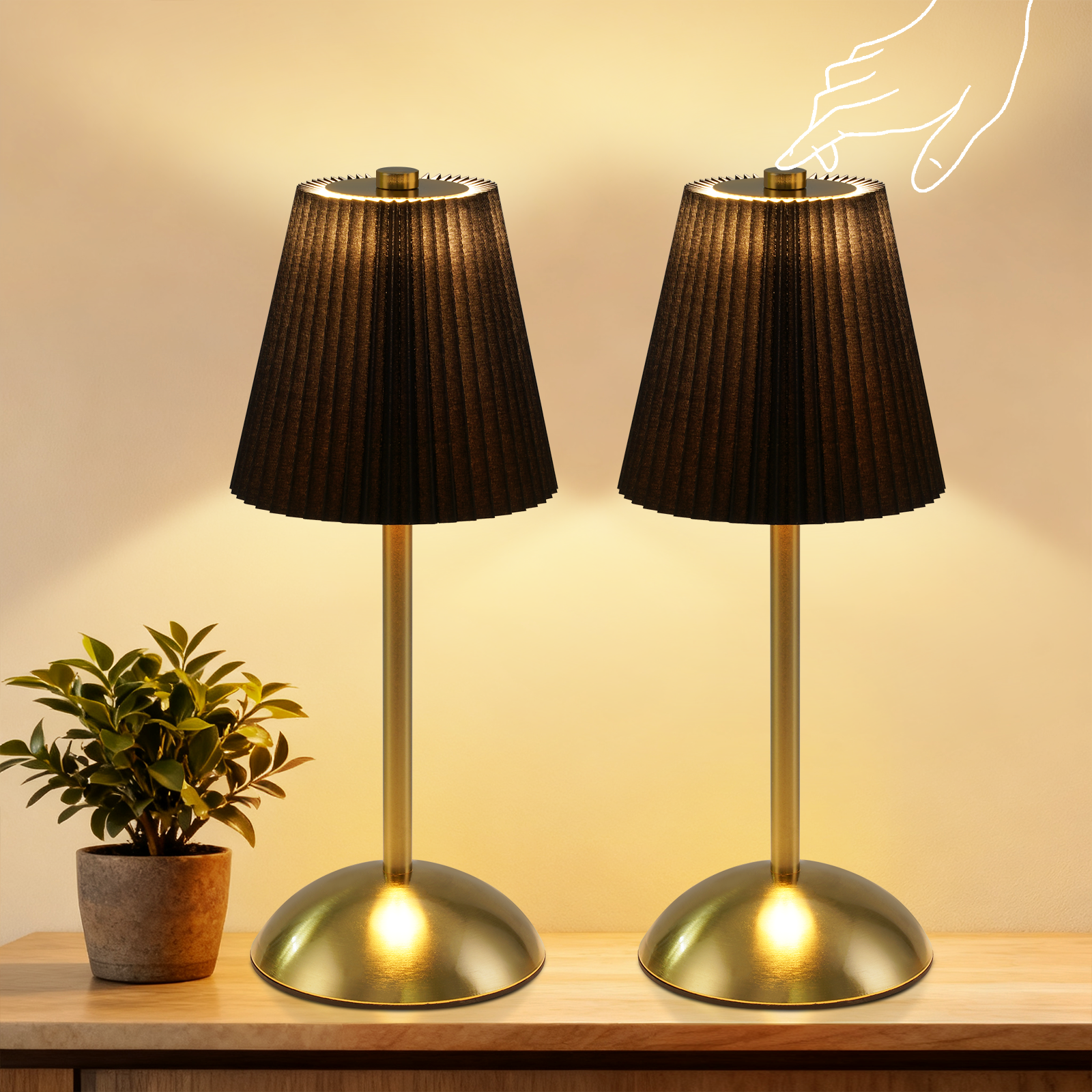 Dwie czarne lampy ze złotymi podstawami na drewnianej powierzchni. Po lewej roślina w doniczce. Ręka ma wcisnąć lampę.