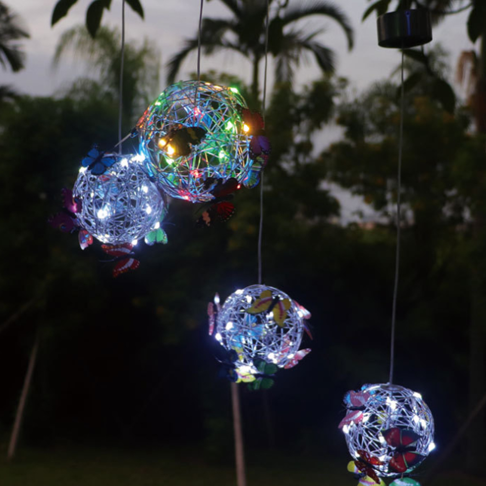 Drei beleuchtete Kugeldekorationen mit Schmetterlingen, die in einem Garten hängen. Die Kugeln haben unterschiedlich farbige Lichter.