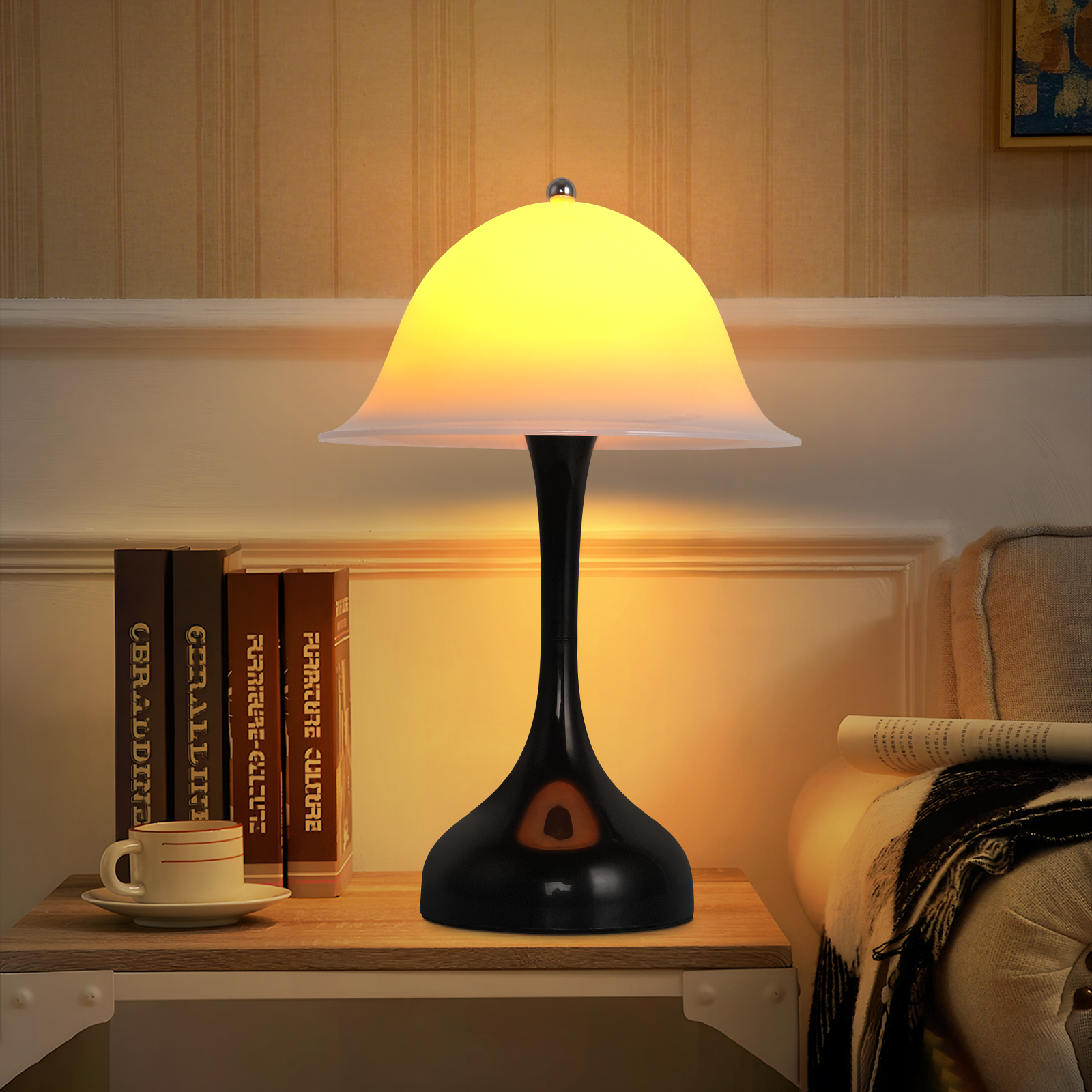 Zapalona lampa stoi na drewnianym stole z książkami i filiżanką kawy. W tle sofa i wzorzysta ściana.