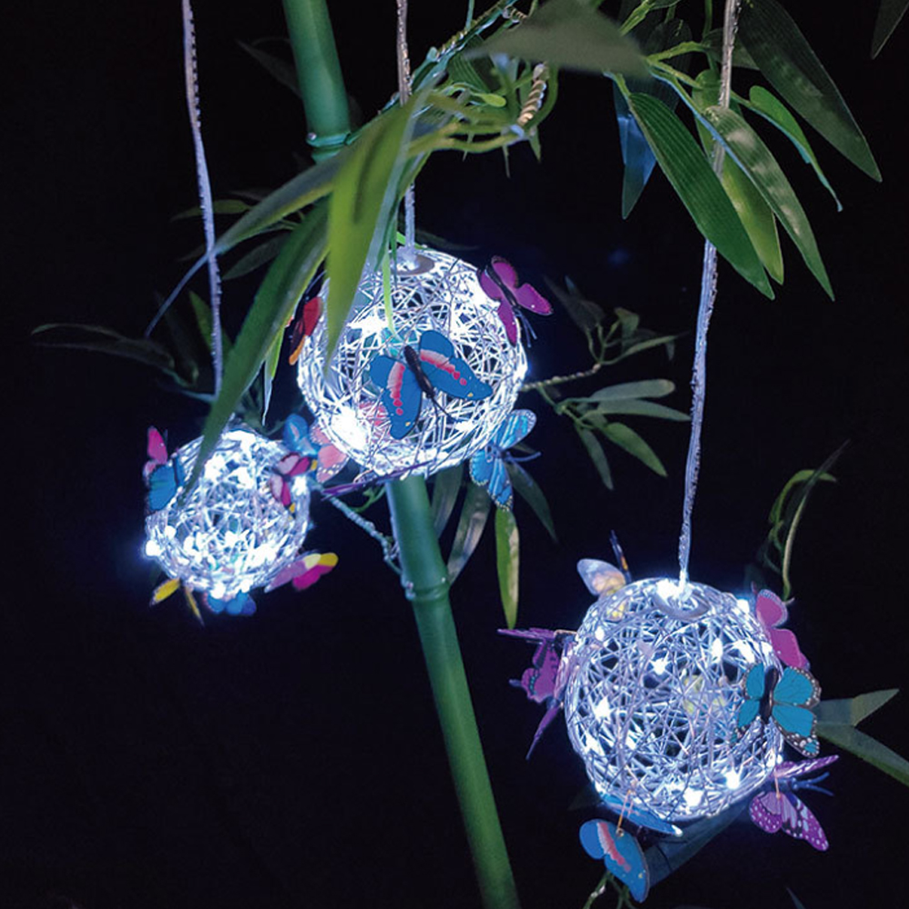 Drei beleuchtete Kugeln, die mit Schmetterlingen verziert sind, hängen an Bambus. Schmetterlinge sind rosa, blau und lila. Dunkler Hintergrund.
