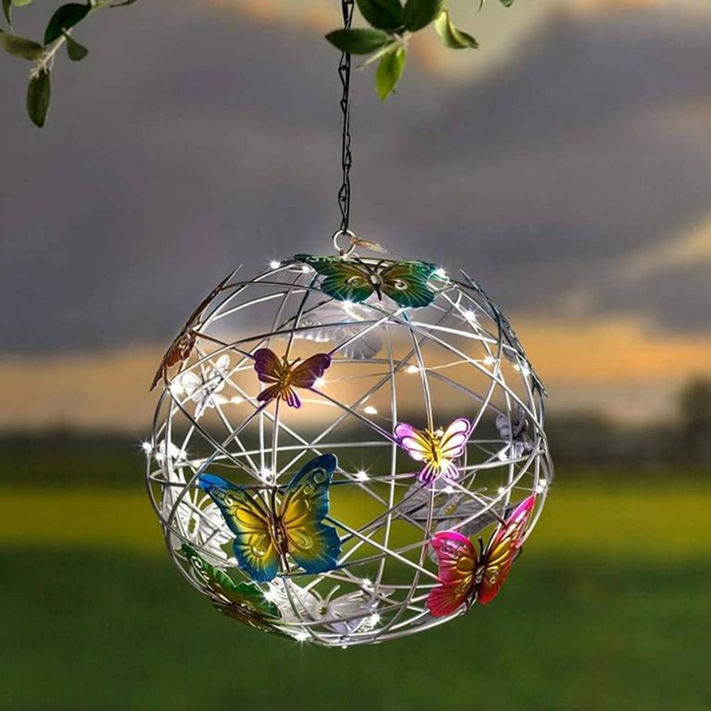 Ein Metallkugel-Ornament mit bunten Schmetterlingsdekorationen, beleuchtet mit kleinen Lichtern, hängt an einem Ast vor einem Sonnenuntergang.