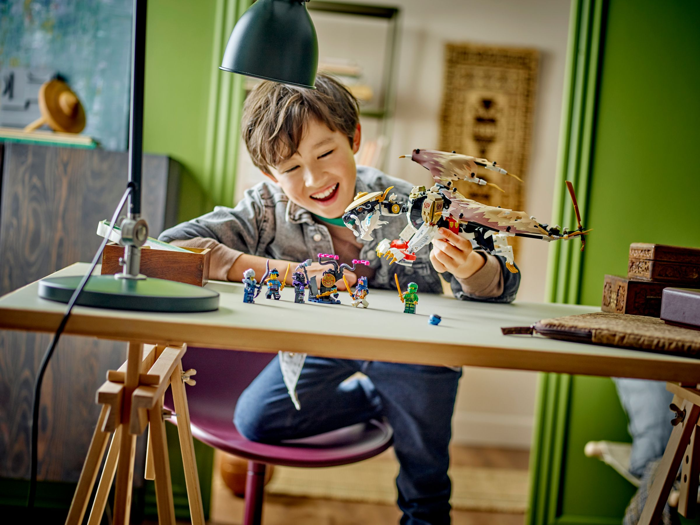 Ein lächelnder Junge hält ein Lego-Set. Das Set enthält einen Drachen und Ninja-Figuren, auf einem Tisch.