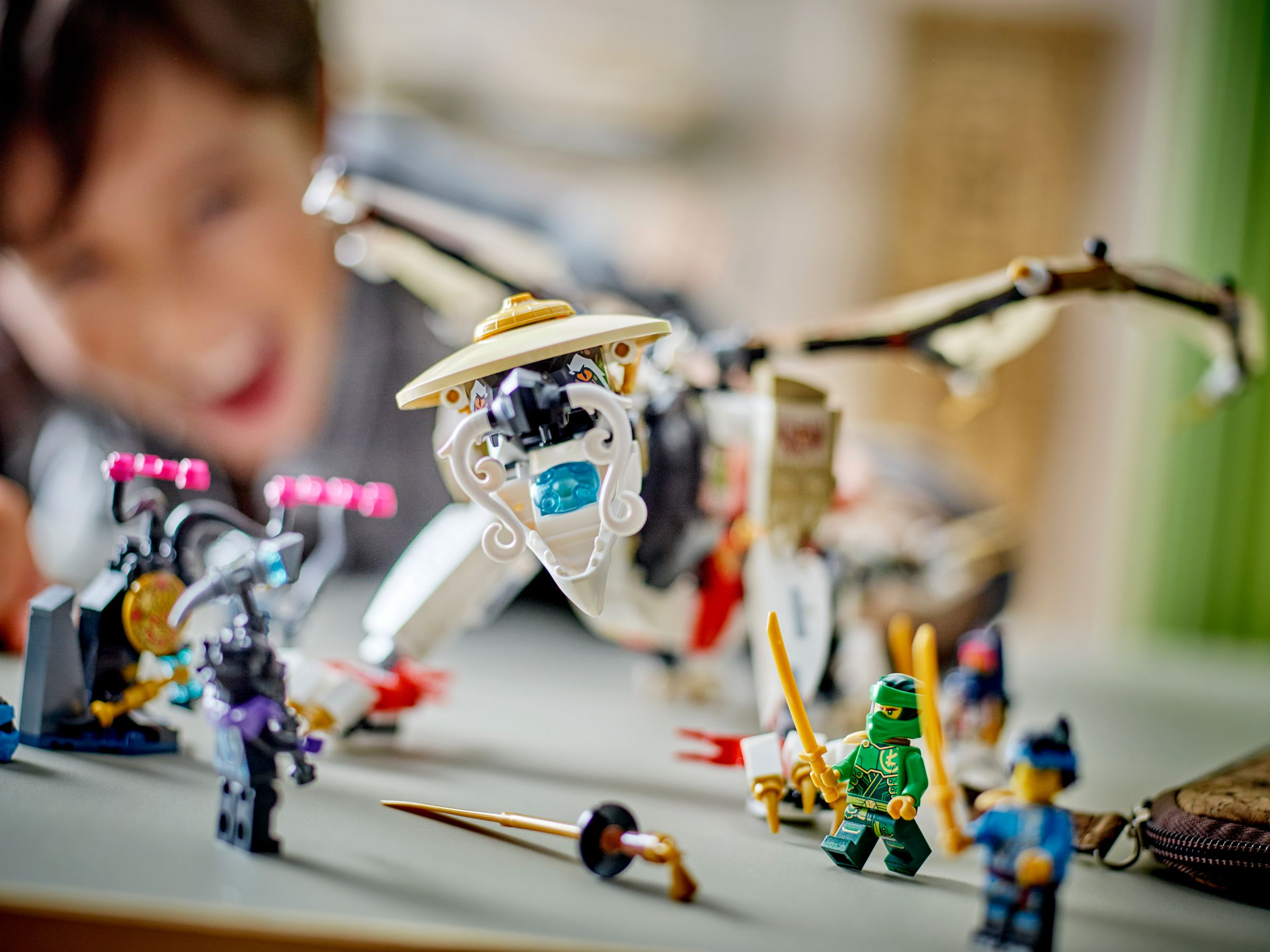 Ein Kind spielt mit einem Lego Ninjago Set, Figuren und einem Drachen. Grüne, gelbe und blaue Figuren. Das Kind ist verschwommen.