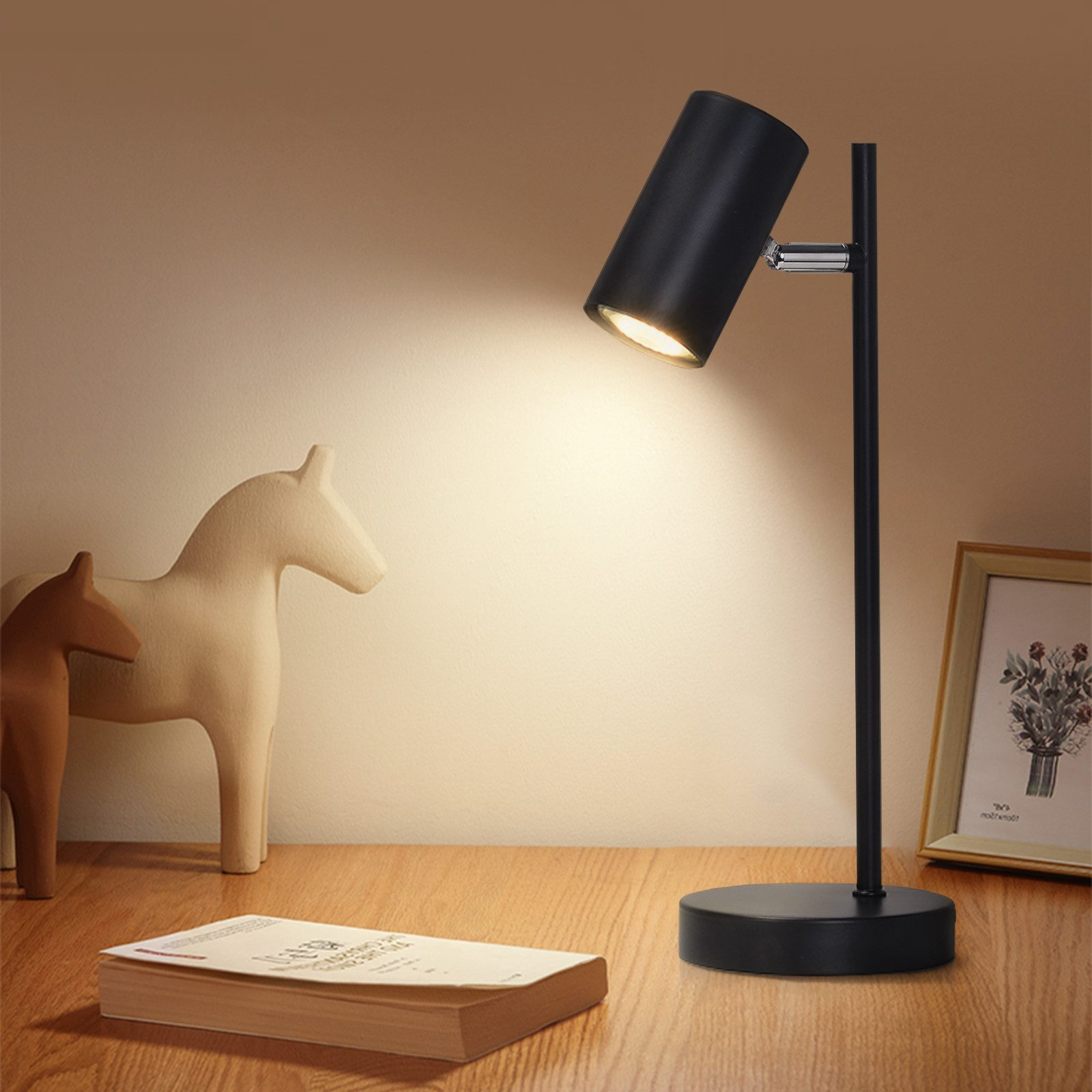 Una lampada da scrivania nera illumina un tavolo di legno. Ci sono statuette di cavalli e un libro. Un'immagine incorniciata è a destra.