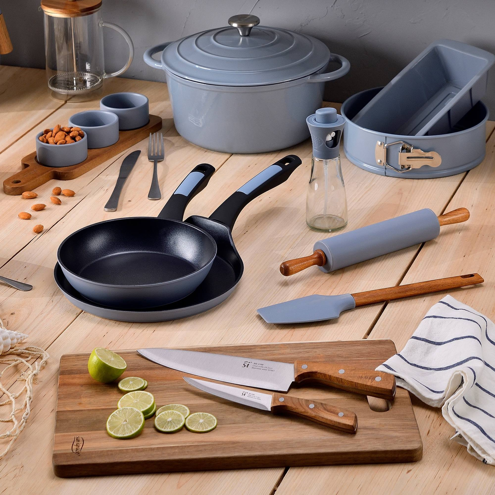 Utensilios de cocina sobre mesa de madera: sartenes, ollas, cuchillos, rodillo, tabla de cortar, ingredientes y cuencos con almendras.