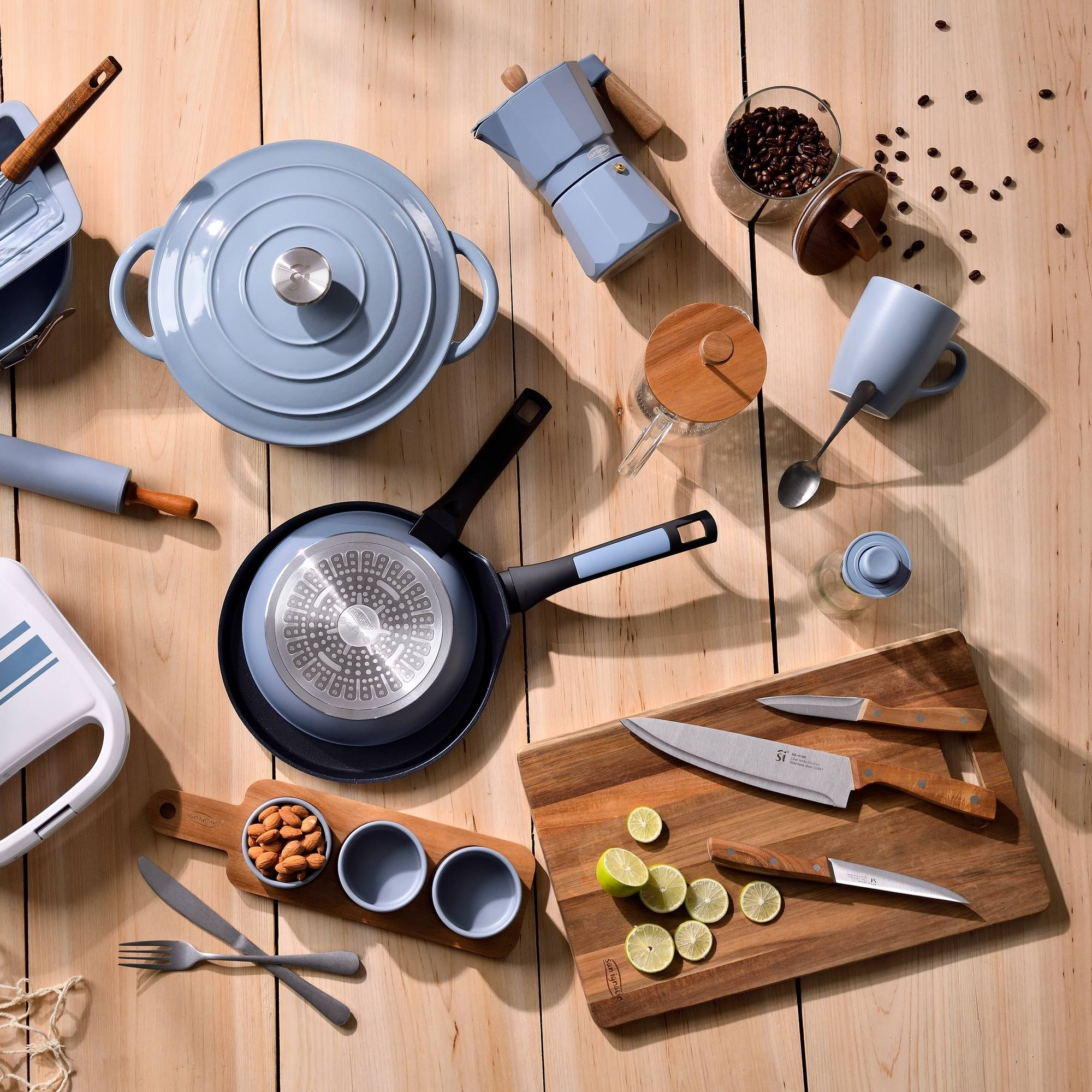 Mesa de madera con utensilios de cocina: olla, sartén, cafetera, cuchillos, granos de café, limones, cuencos, tenedor, cuchillo.