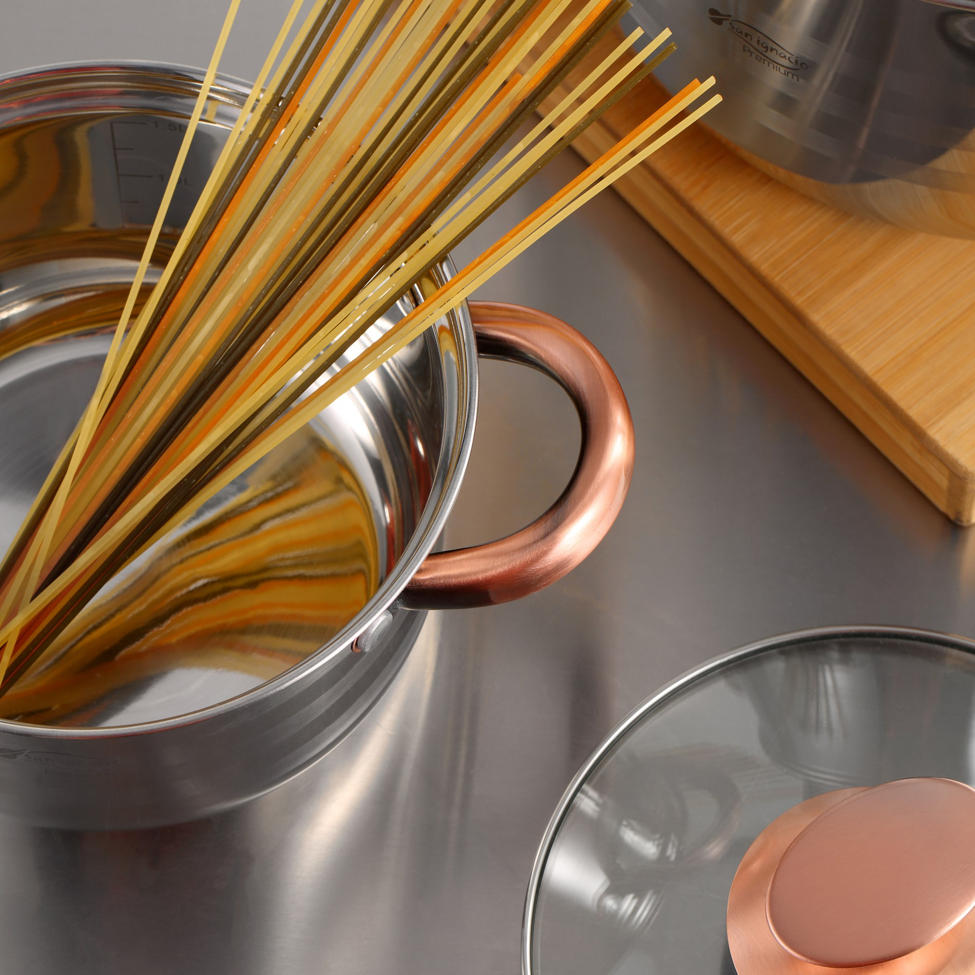Espaguetis en una olla de acero inoxidable con asa de cobre. Tapa en primer plano, tabla de cortar de madera al fondo.