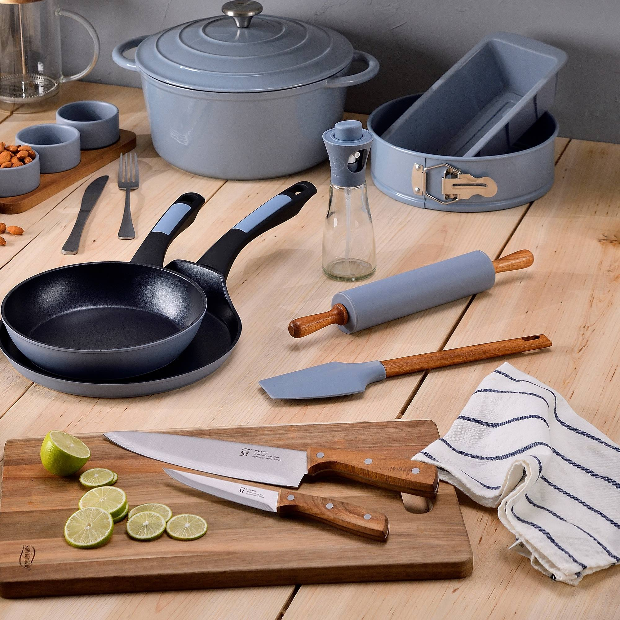 Utensilios de cocina sobre una mesa de madera: ollas, sartenes, cuchillos, rodillo y tabla de cortar con rodajas de lima.
