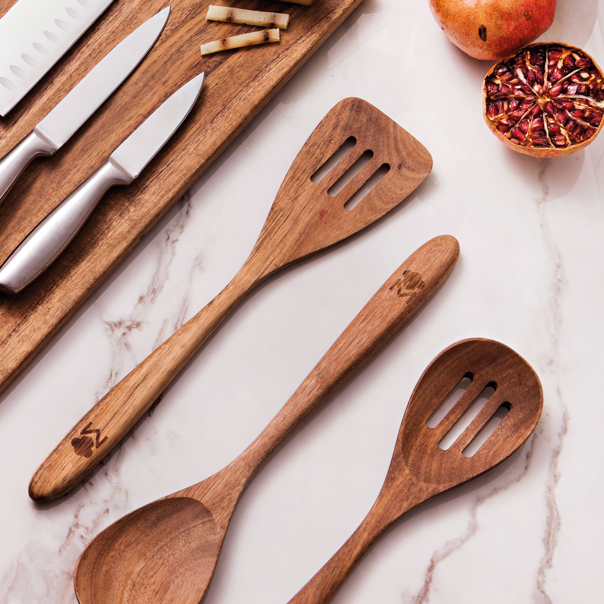 Utensilios de cocina de madera con cuchillos y una granada sobre una superficie de mármol. La disposición es cenital.