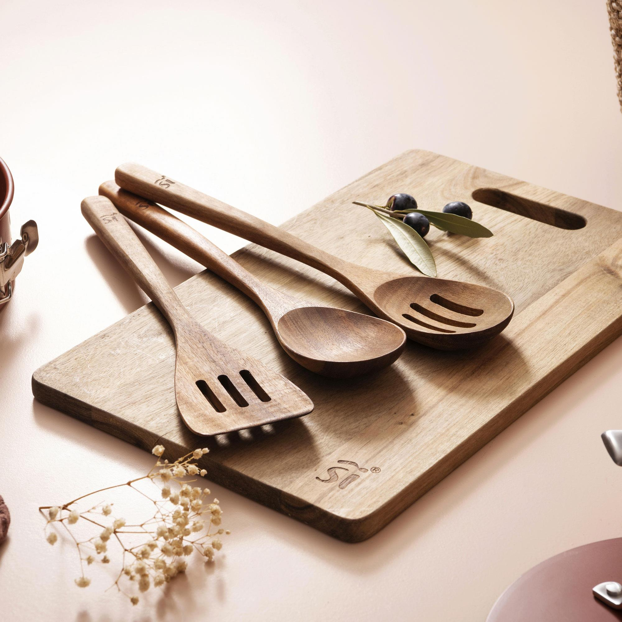 Utensilios de cocina de madera sobre una tabla de madera con aceitunas. Incluye espátula, cuchara y cuchara ranurada. Fondo rosa suave.